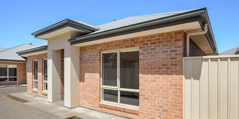 Front exterior view of a brick house with large windows, a small covered entryway, a concrete sidewalk, and a beige metal fence in the yard, under a clear blue sky.