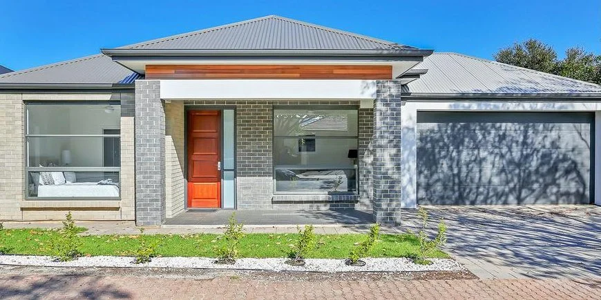 Modern house with brick and wood exterior, featuring a red front door, large front window, and an attached garage, with a small garden in front.