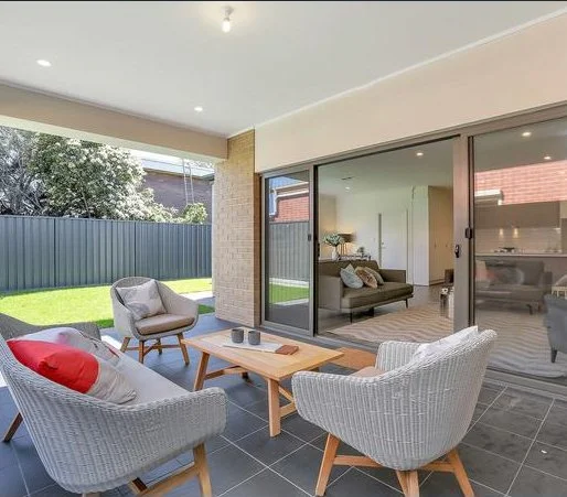 Outdoor patio with four white wicker chairs and a wooden coffee table, overlooking a green backyard with a metal fence and trees visible in the background.