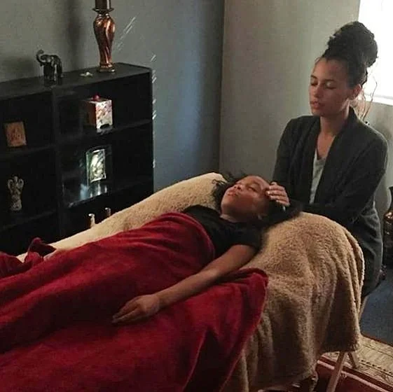 A woman is giving reiki to a person lying on a massage table covered with red and beige blankets in a room with dark-colored walls and a black shelf with decorative items.