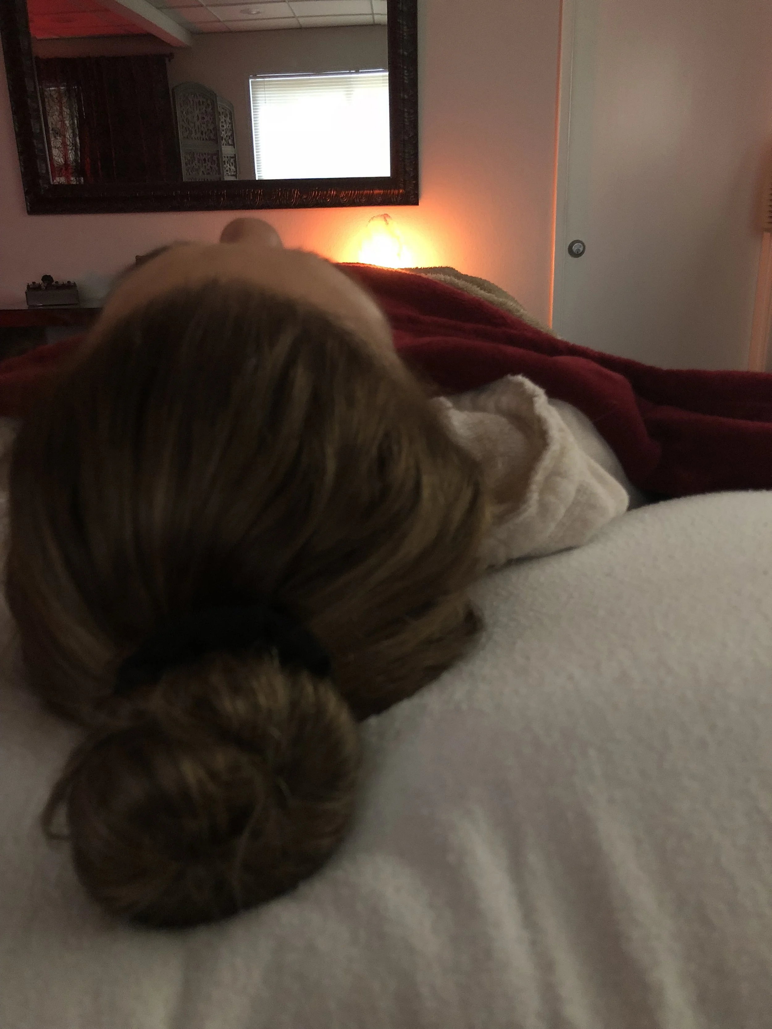 A person lying on a bed resting on their back, in a room with a mirror, window, and some furniture in the background.