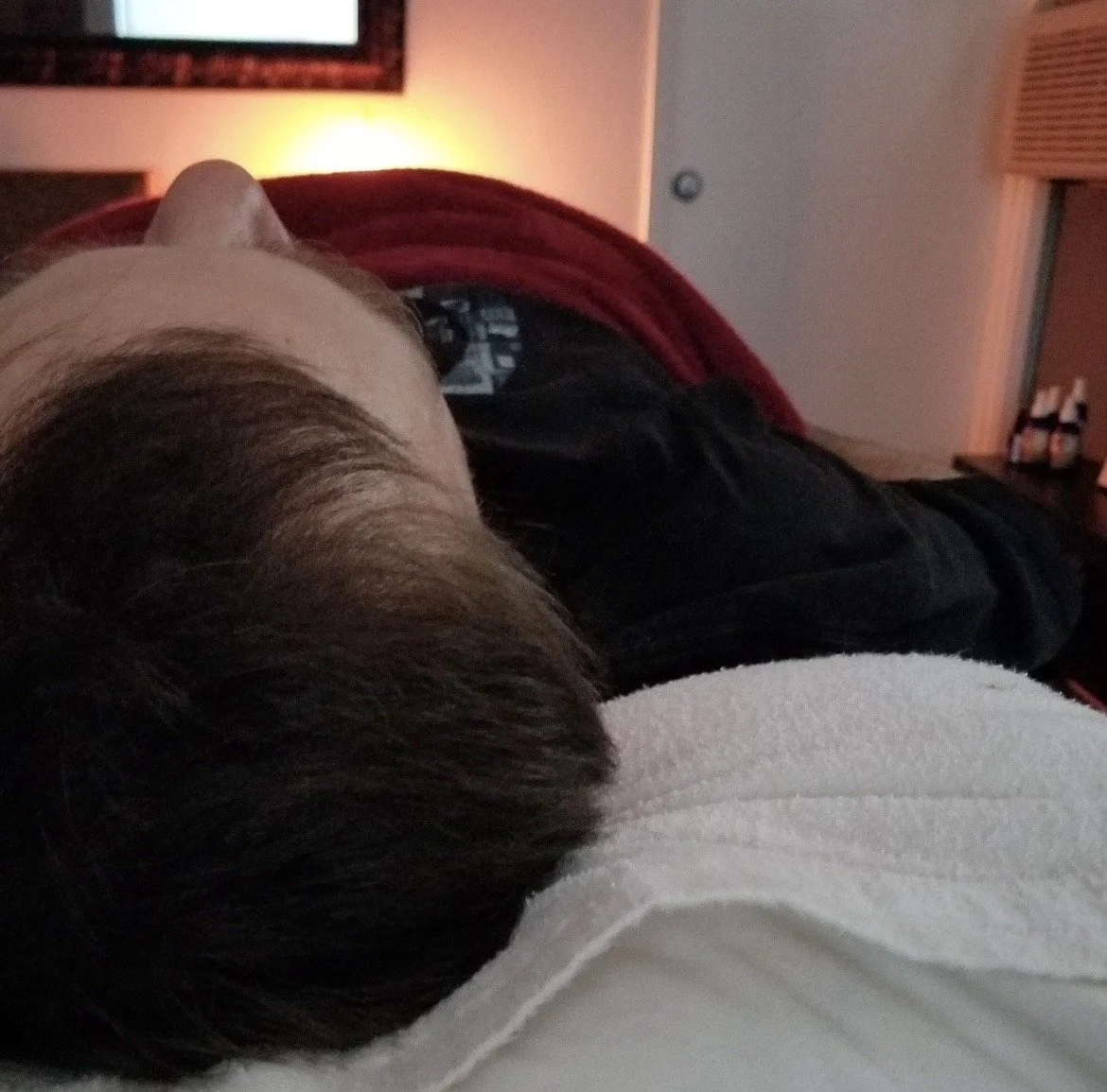 Person lying down on a white pillow with their head turned to the side in a dimly lit room, with a television and some bottles visible in the background.