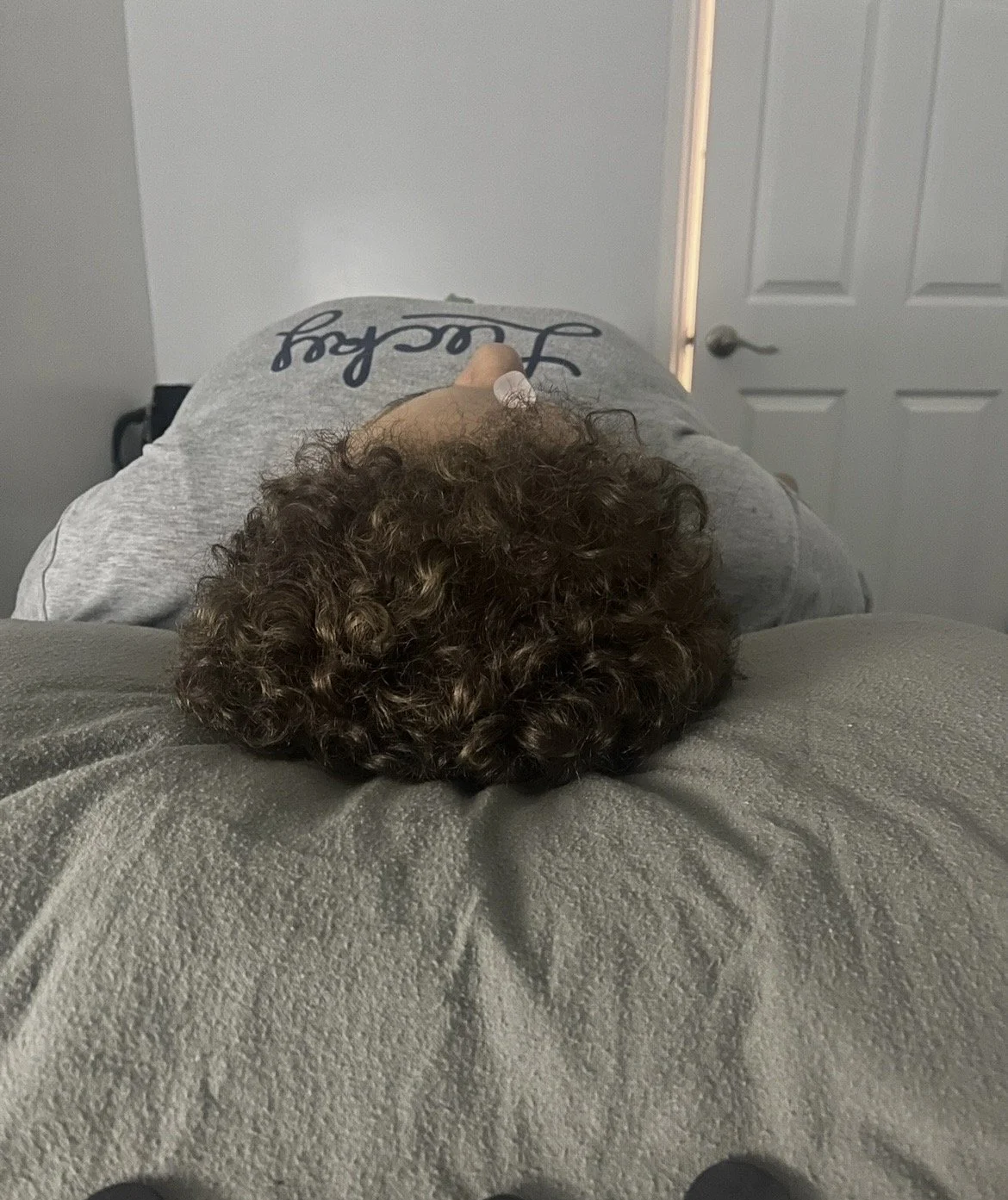 A person with curly hair lying face down on a bed, wearing a gray shirt with dark text, in a room with a closed white door in the background.