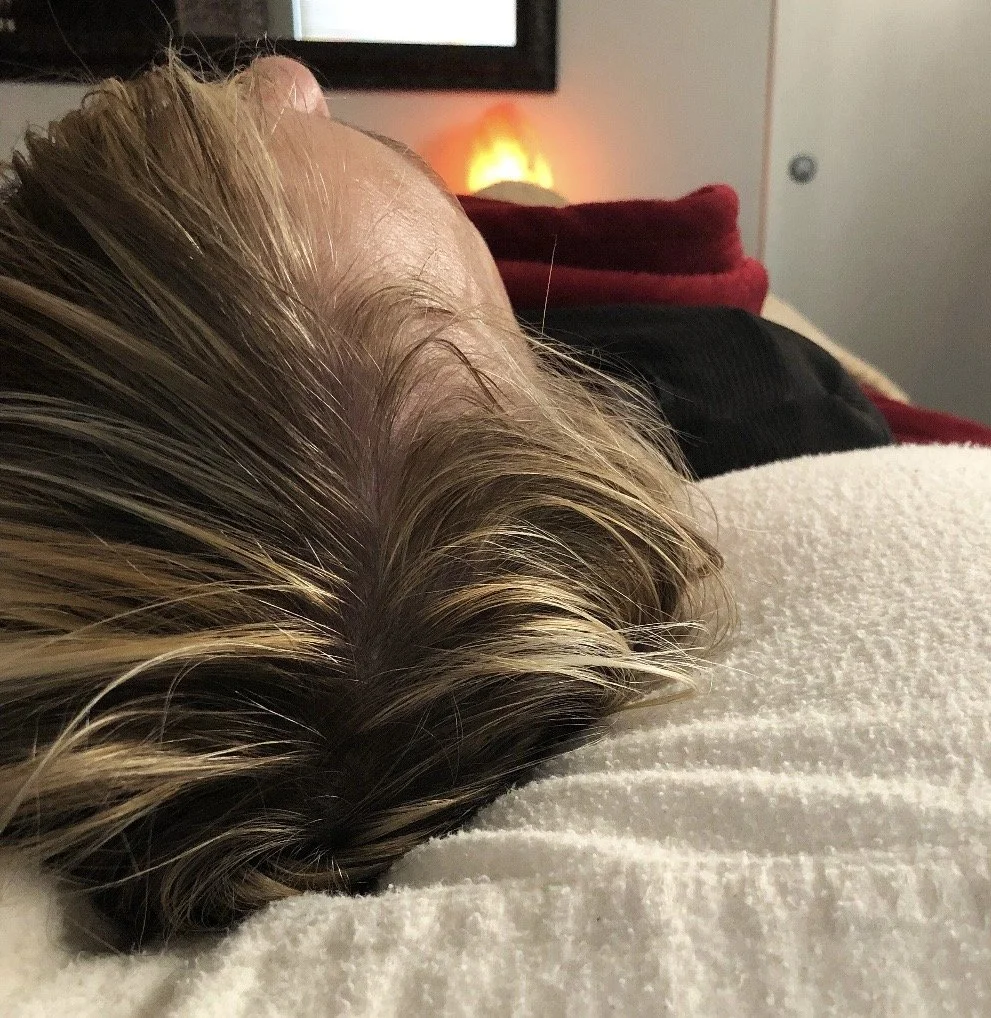 A person with blonde hair lying on a white soft surface, possibly a bed or couch, with a red blanket and a black piece of clothing, in a cozy room with a lit Himalayan salt lamp and a television in the background.