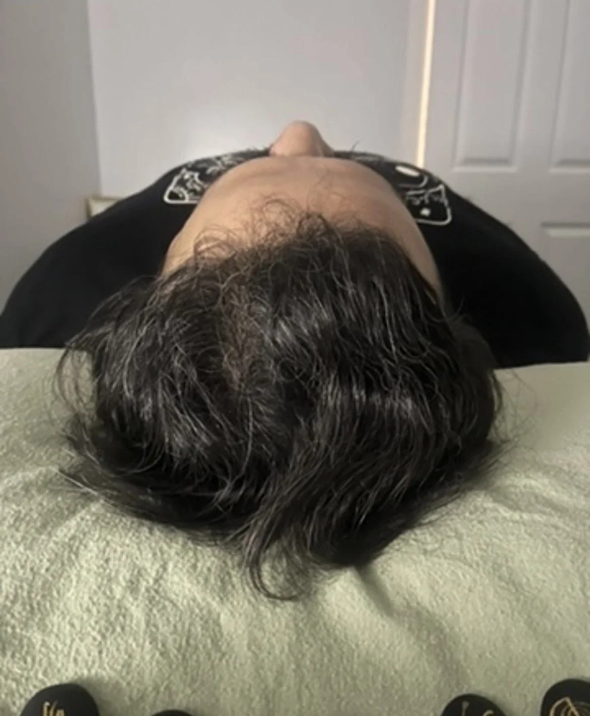 Top view of a person lying on a surface with their head facing down, showing dark hair with some gray strands.