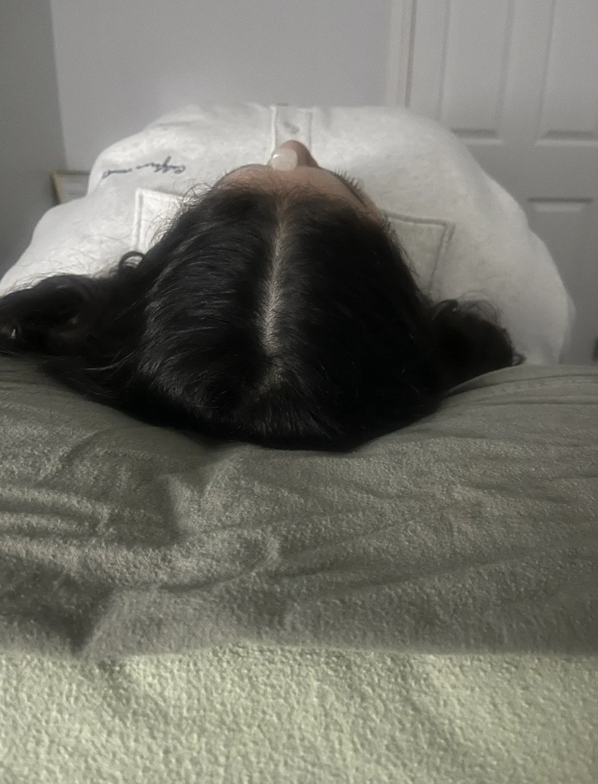 A person lying on their back on a bed with dark hair parted in the middle, wearing a white shirt, and crystal on their head.