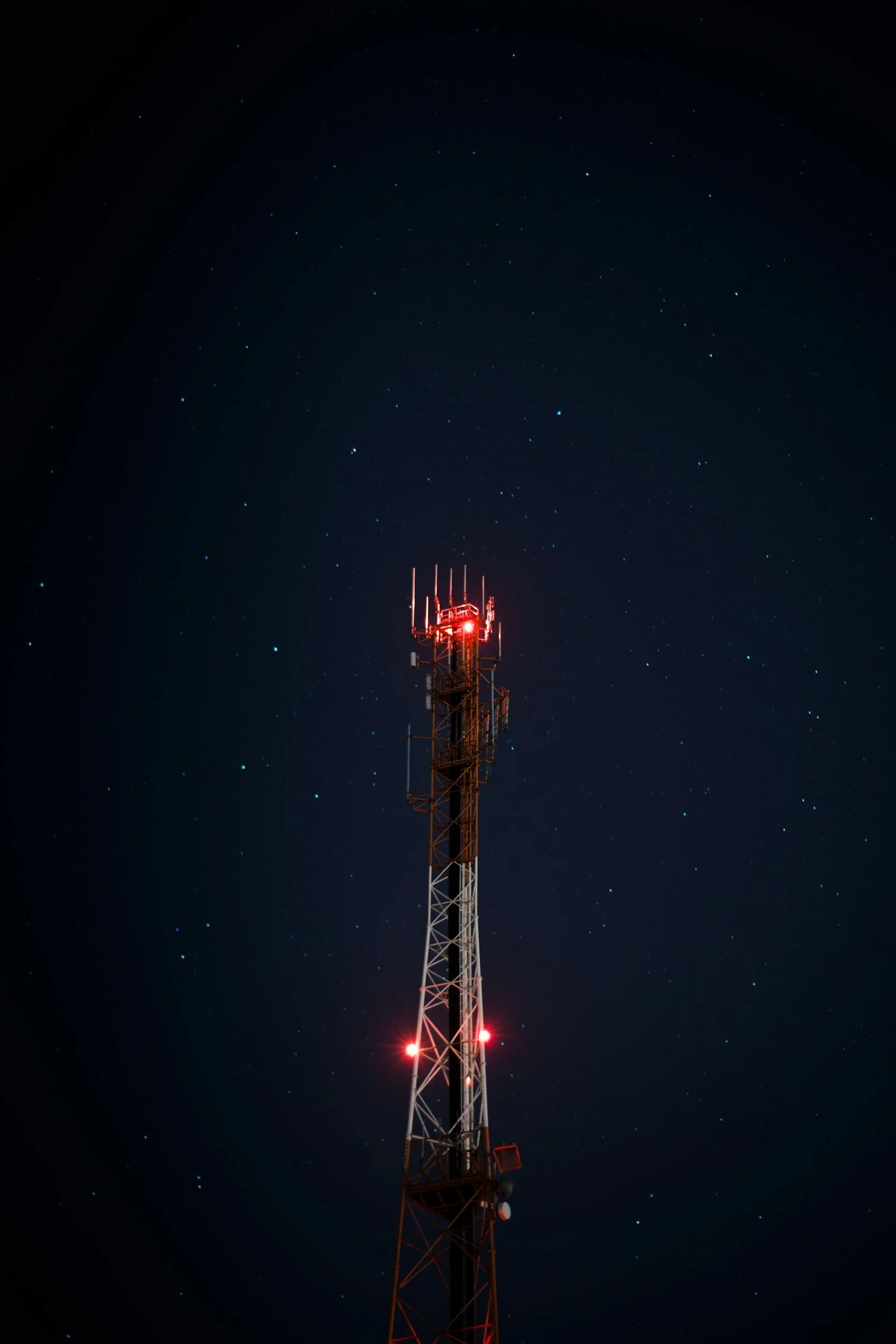 Night sky filled with stars above a tall telecommunications tower with red warning lights.