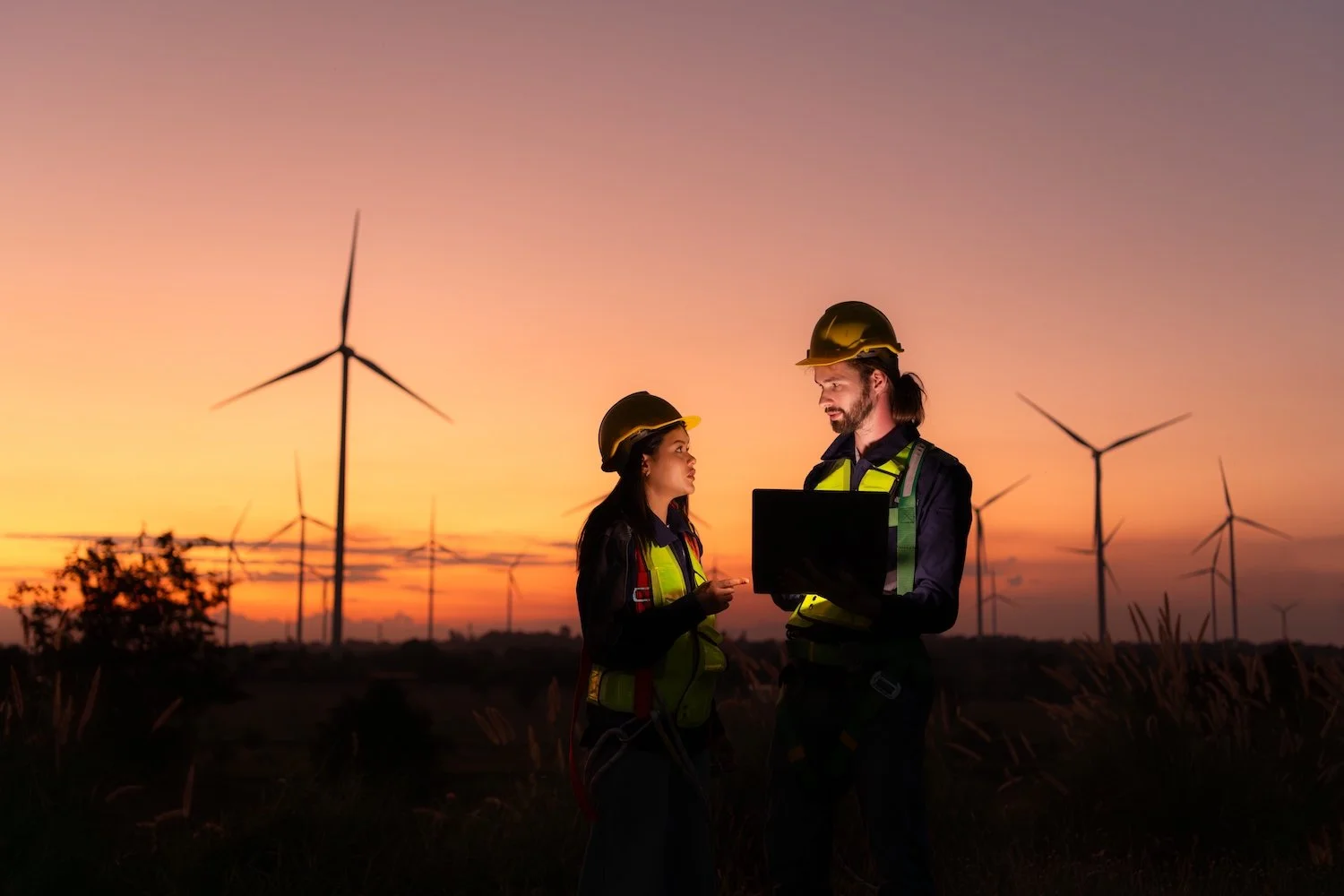 Two wind turbine technicians in safety gear discuss work on wind turbines at sunset, holding a laptop, in a field.