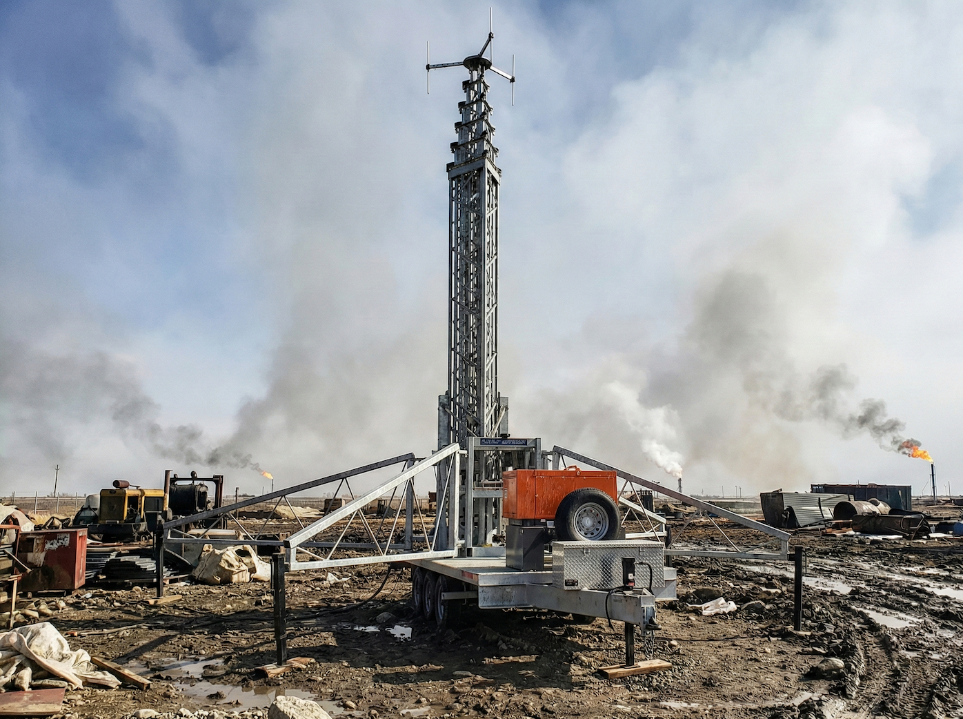 An oil drill rig in a barren landscape with smoke stacks in the background and oil fires burning in the distance.
