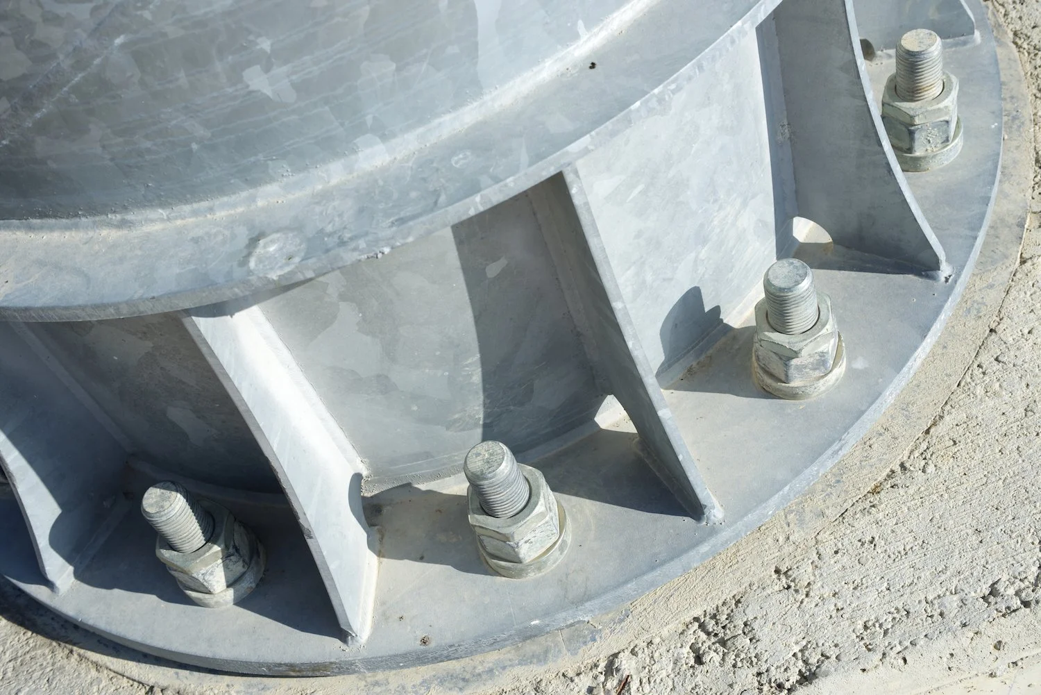 Close-up of a metal base with large bolts securing it to the ground. The base has a curved, circular shape with support brackets, and is positioned on concrete ground.