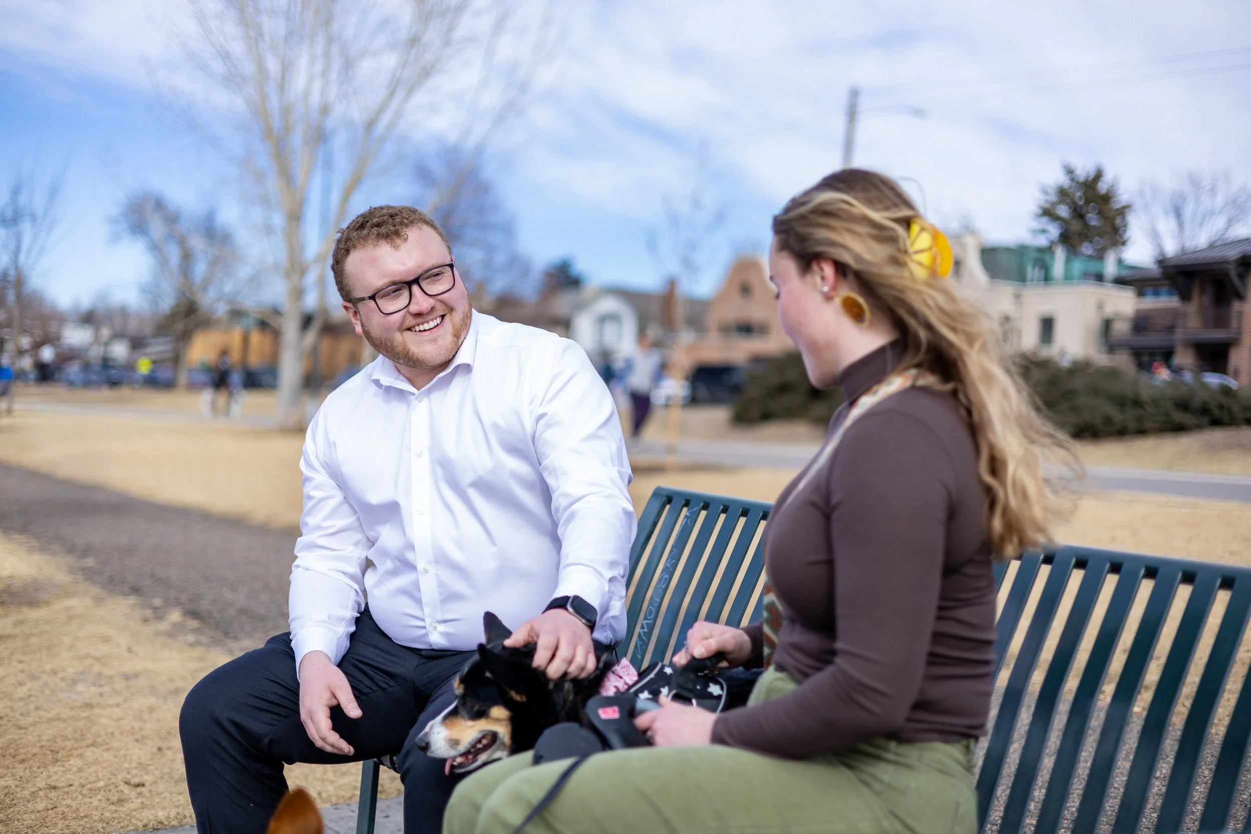 Matt Walter and a woman sitting on a park bench, smiling and engaging in conversation, with a small dog on the woman's lap during daytime.