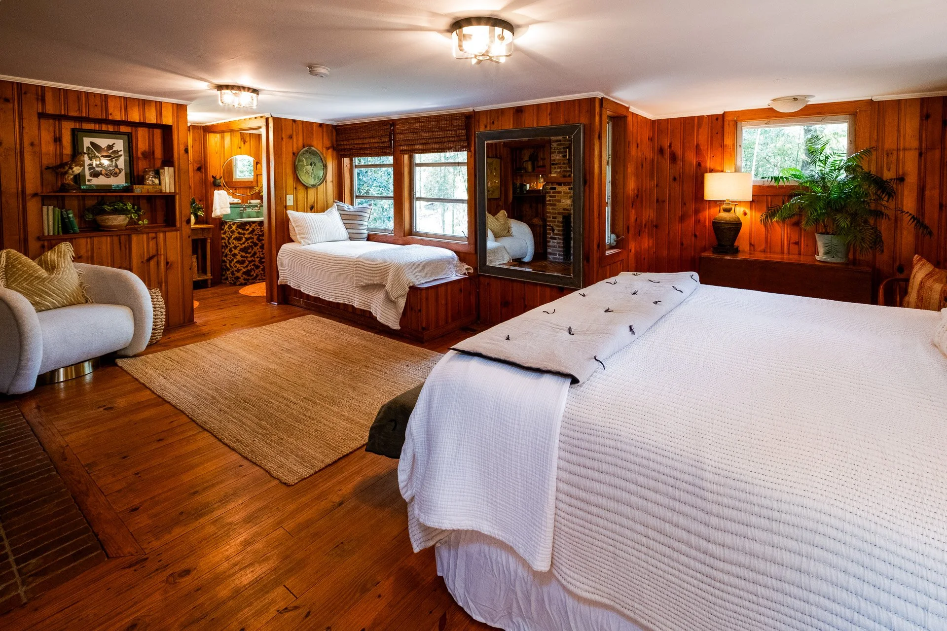 A cozy bedroom with wood-paneled walls, a large bed with white bedding, a sitting area with a beige armchair, a window seat, and a small bedroom area with a single bed. There is a rug on the wooden floor, plants, and lamps providing warm lighting.