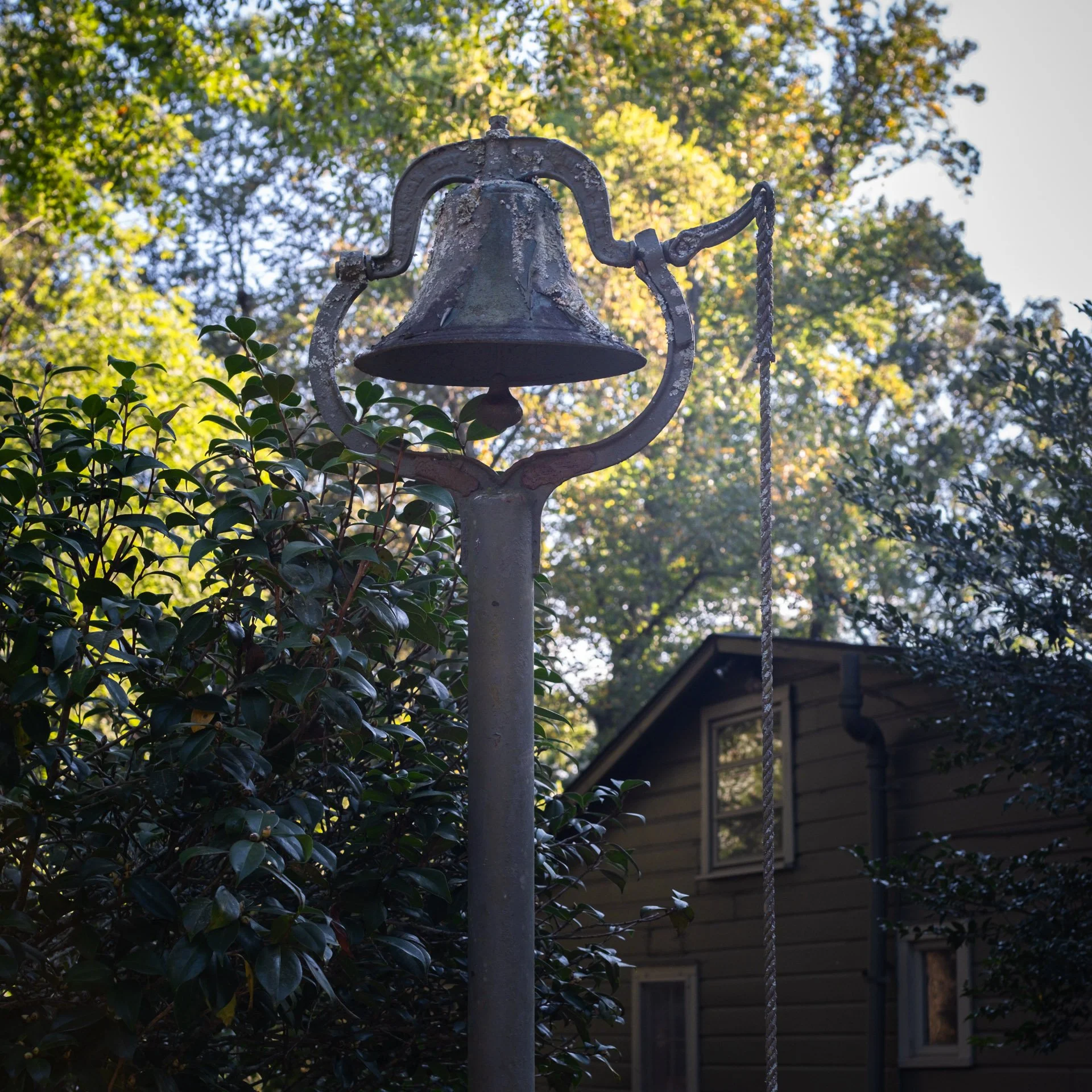 An old metal outdoor bell attached to a pole, with a rope for ringing, set against trees and a house in the background.