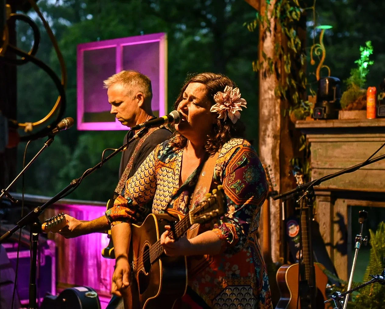 Two musicians performing outdoors at dusk; one woman singing and playing an acoustic guitar with a flower in her hair, one man playing an electric guitar beside her, set against decorated natural surroundings.