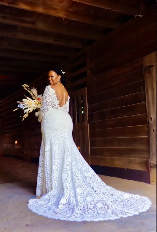 A bride in a white lace wedding gown standing indoors with wooden walls, holding a bouquet of flowers, and smiling while looking over her shoulder.