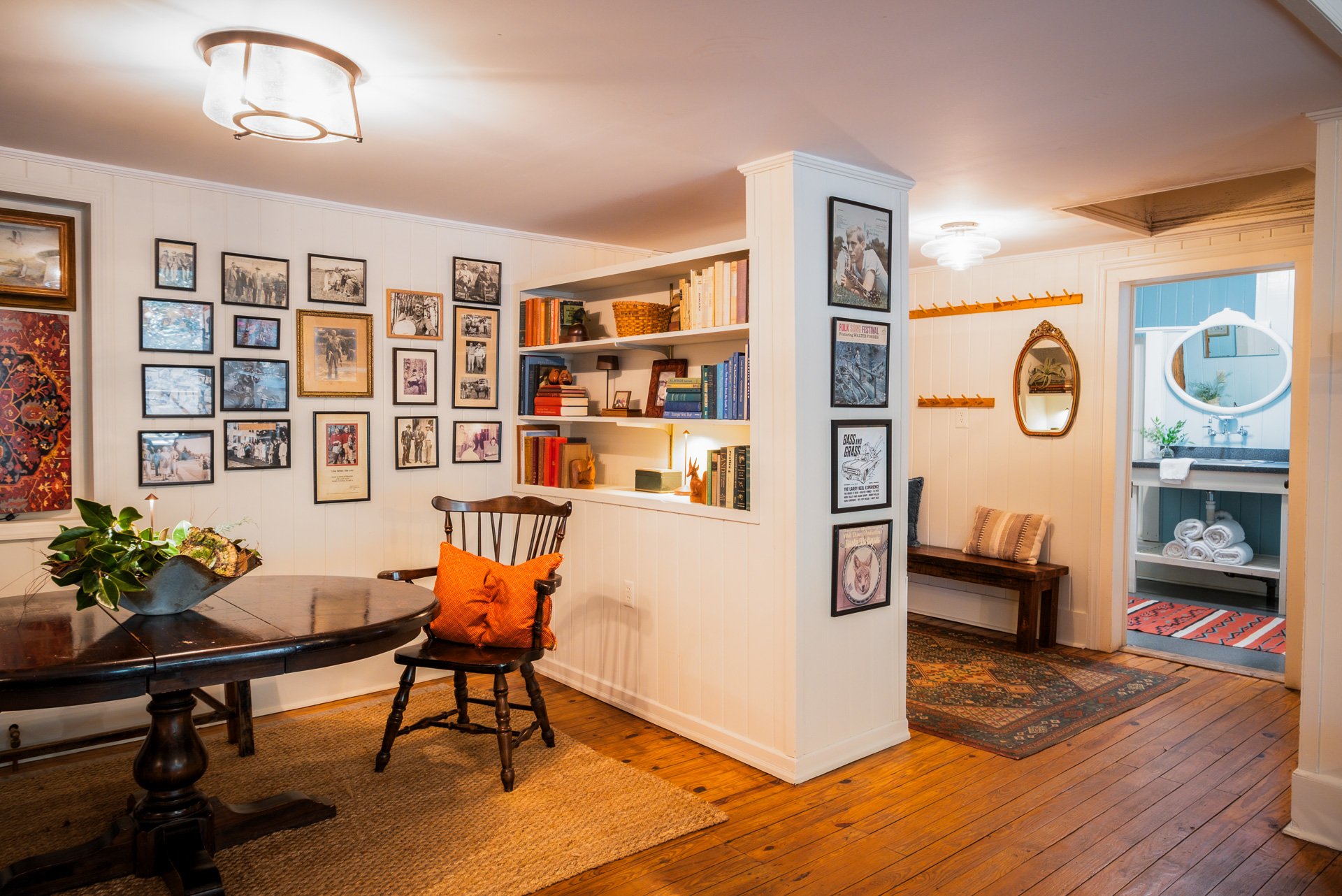 A cozy home interior with a corner bookshelf, a gallery wall of photos, a dining table with a wooden chair featuring an orange pillow, a small hallway leading to a bathroom with rolled towels, and wooden flooring.