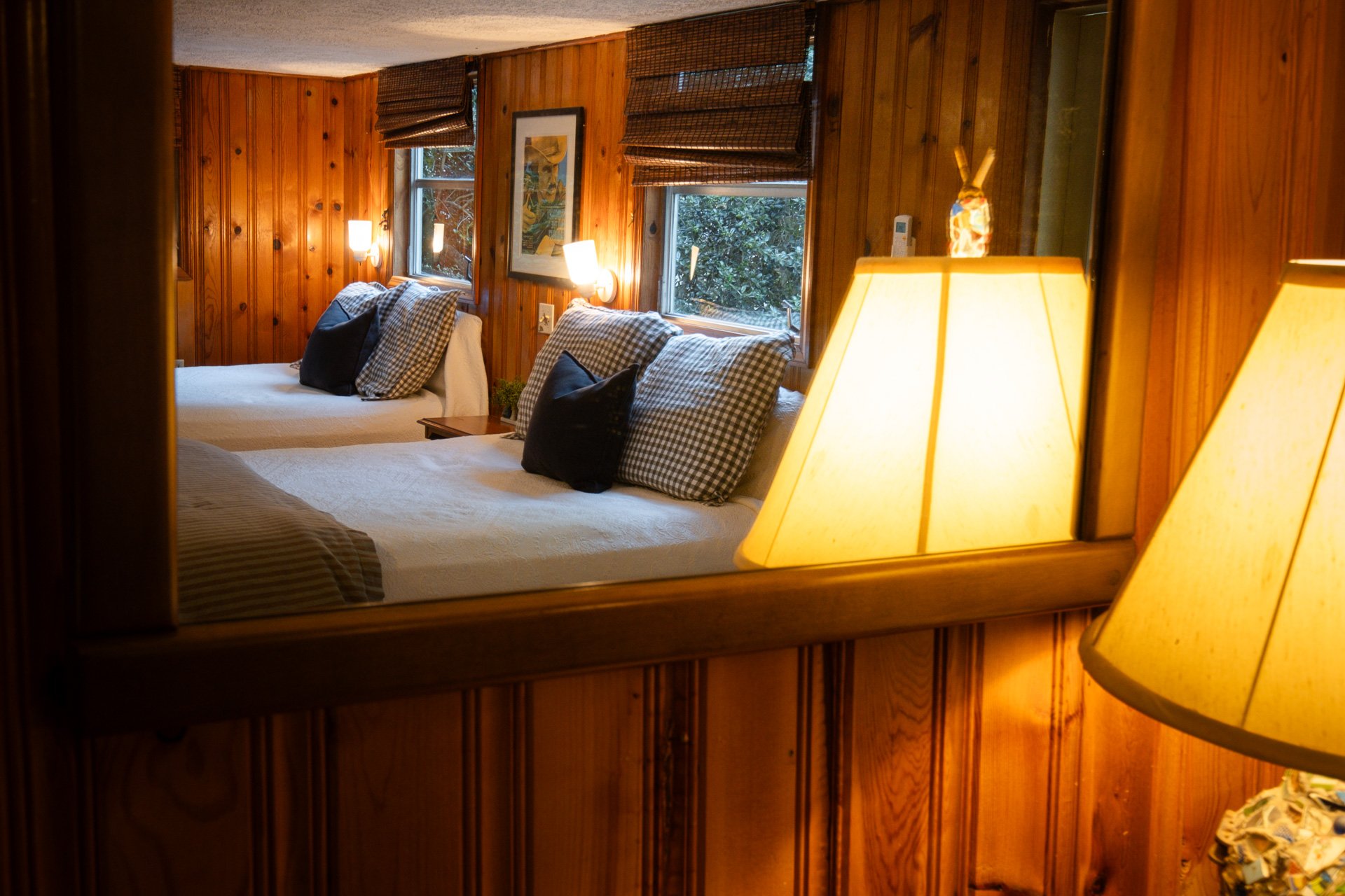A cozy bedroom with wooden walls and ceiling, featuring two beds with plaid and black pillows, multiple windows with bamboo blinds, and warm lighting from lamps and wall-mounted fixtures.
