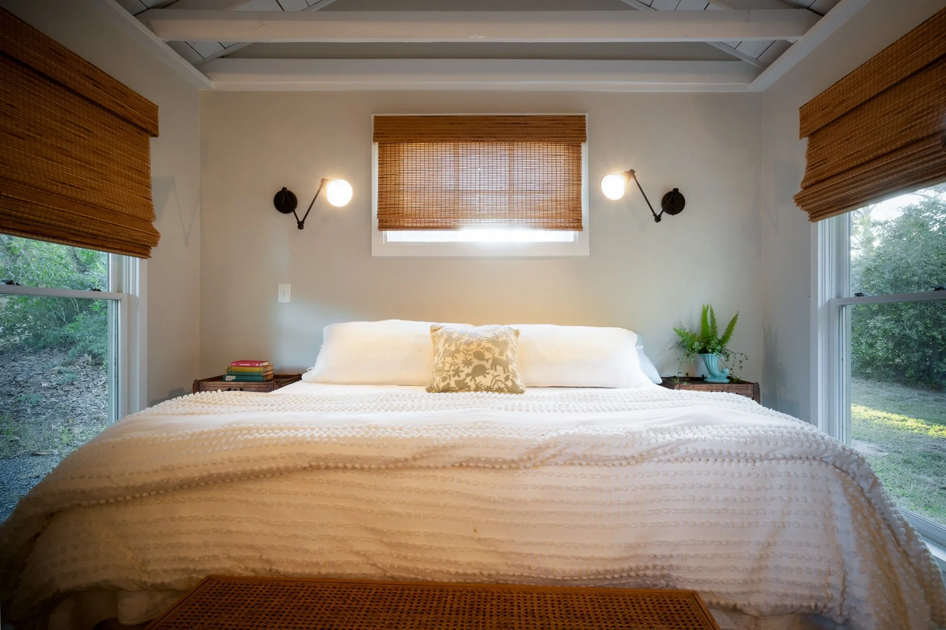 A cozy bedroom with a large bed covered in a white textured blanket, a decorative pillow, wooden side tables with books, a potted plant, and windows with bamboo shades, lit by wall-mounted lamps.