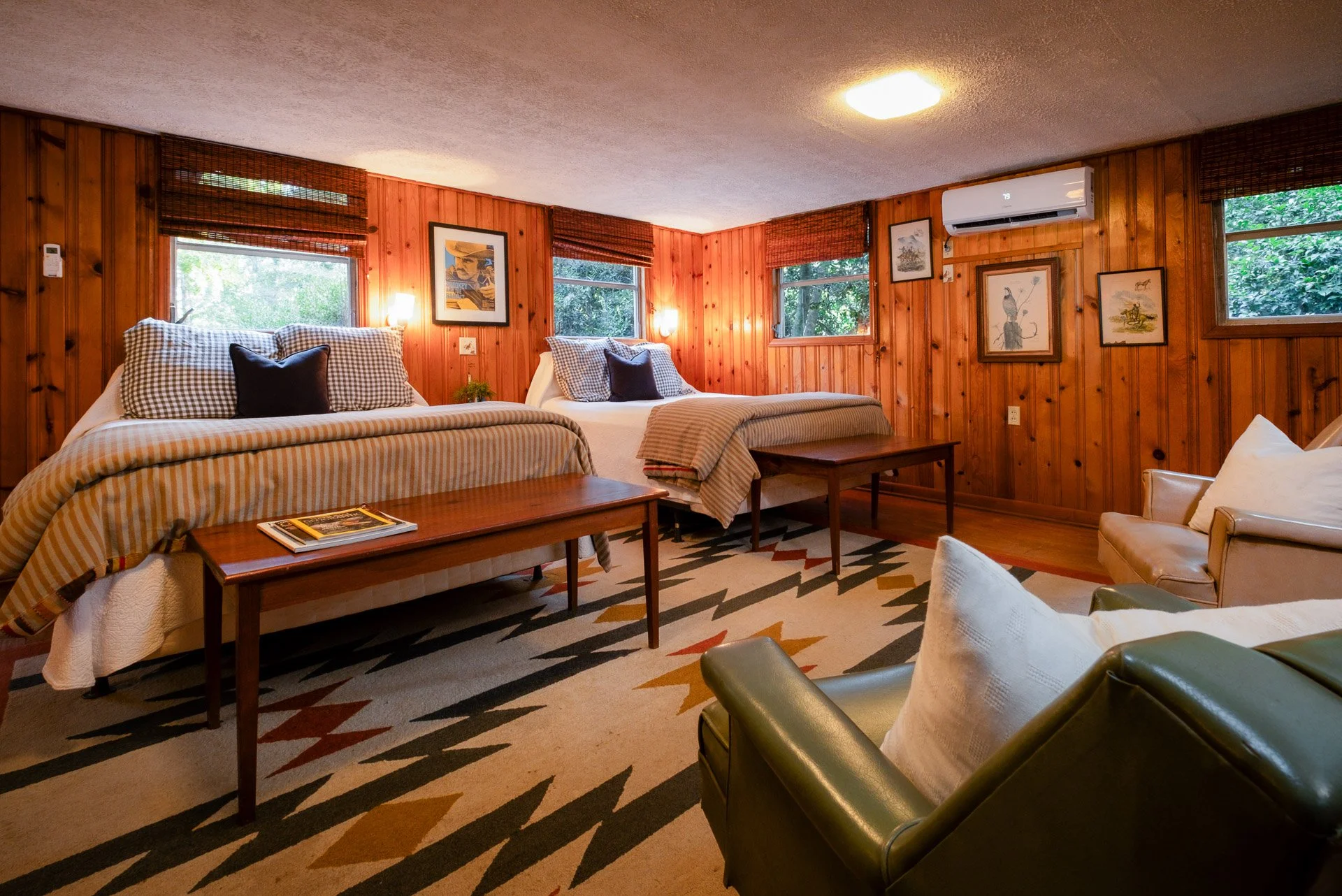 A cozy wooden-paneled hotel room with two large beds, each made up with striped bedding and Checkered pillows, a small cream-colored armchair, and a couch with pillows. There are three windows showing greenery outside, framed by wooden blinds. Wall art features various nature illustrations. A patterned rug covers the floor, and there are wooden benches at the foot of each bed.