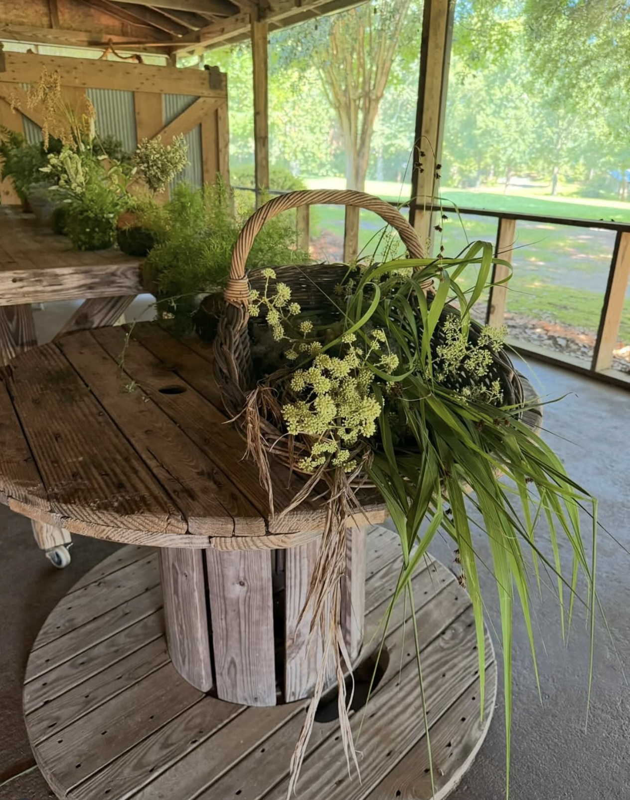 A wicker basket filled with greenery and small white flowers placed on a rustic wooden table.