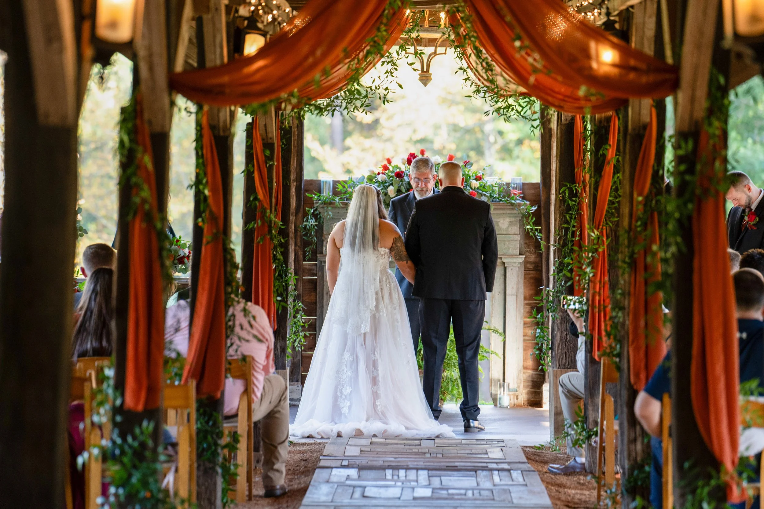 A couple getting married at an outdoor rustic wedding ceremony, with an officiant and guests seated, decorated with orange drapes and greenery.