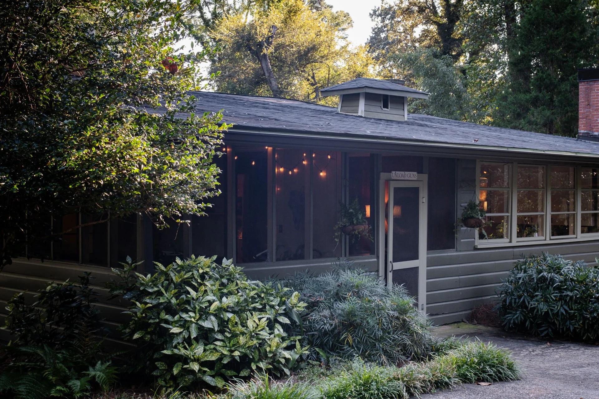A house exterior with a screened porch surrounded by trees and bushes, with string lights hanging inside and a sign on the door that reads 'UNLOAD GUNS'.