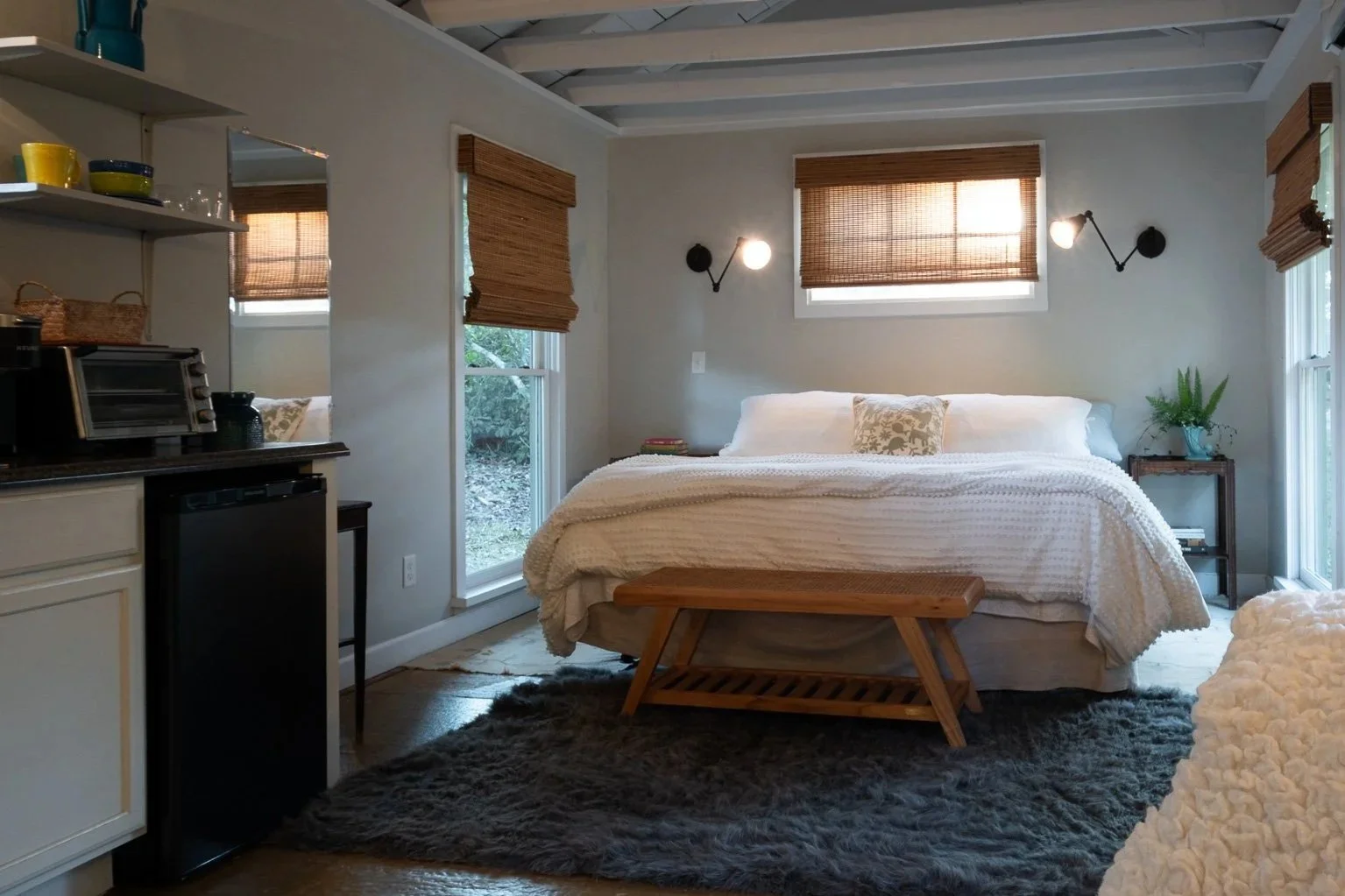 A cozy bedroom with a white bed, wooden bench, and side table, illuminated by natural light from windows with bamboo blinds, and accented by wall-mounted lamps, textured rug, and houseplants.