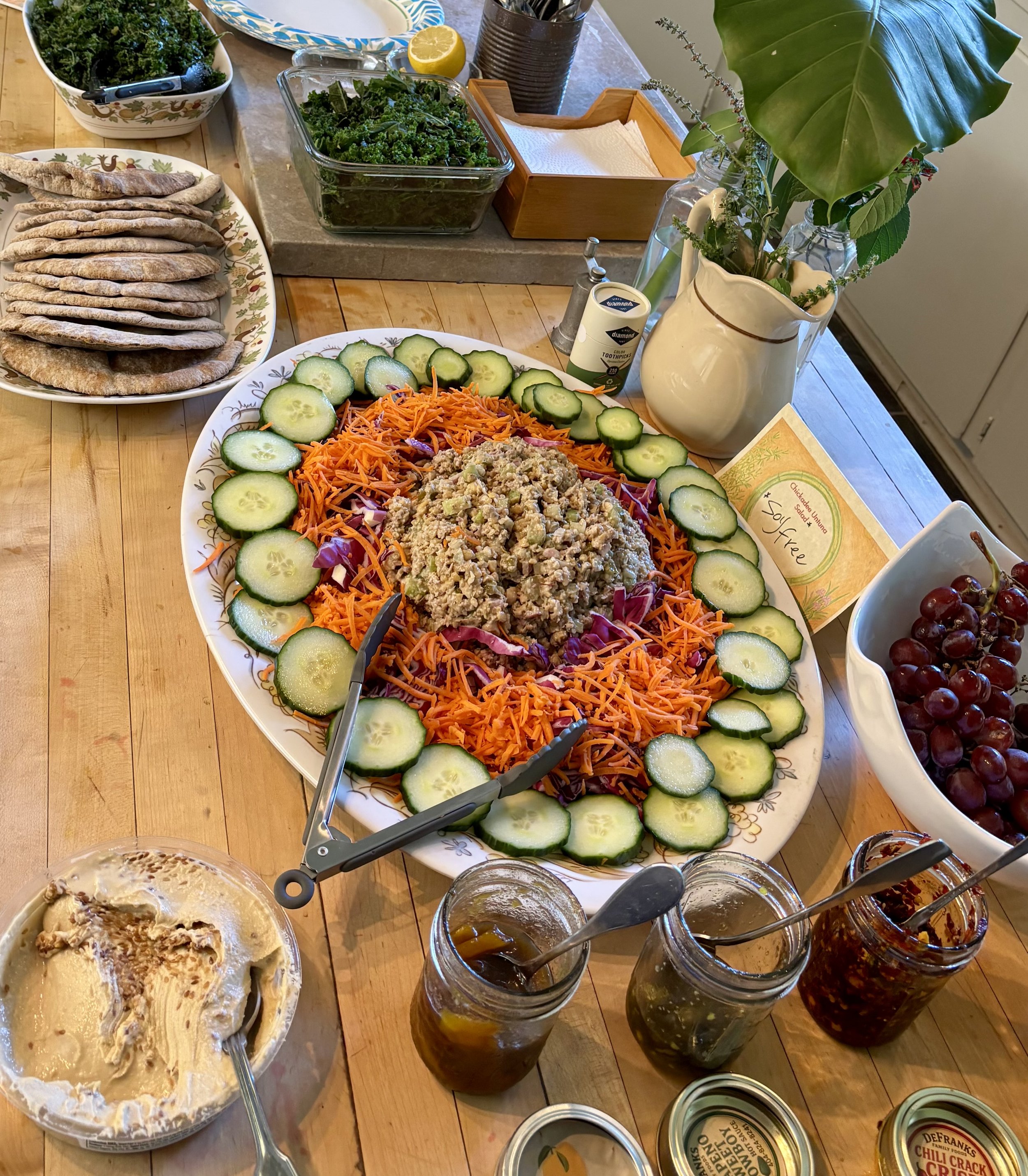 A table with a large vegetable salad featuring shredded carrots, cucumber slices, purple cabbage, and a scoop of chickpea salad in the center. Surrounding the salad are various dishes including a bowl of red grapes, a bowl of pita bread, multiple jar