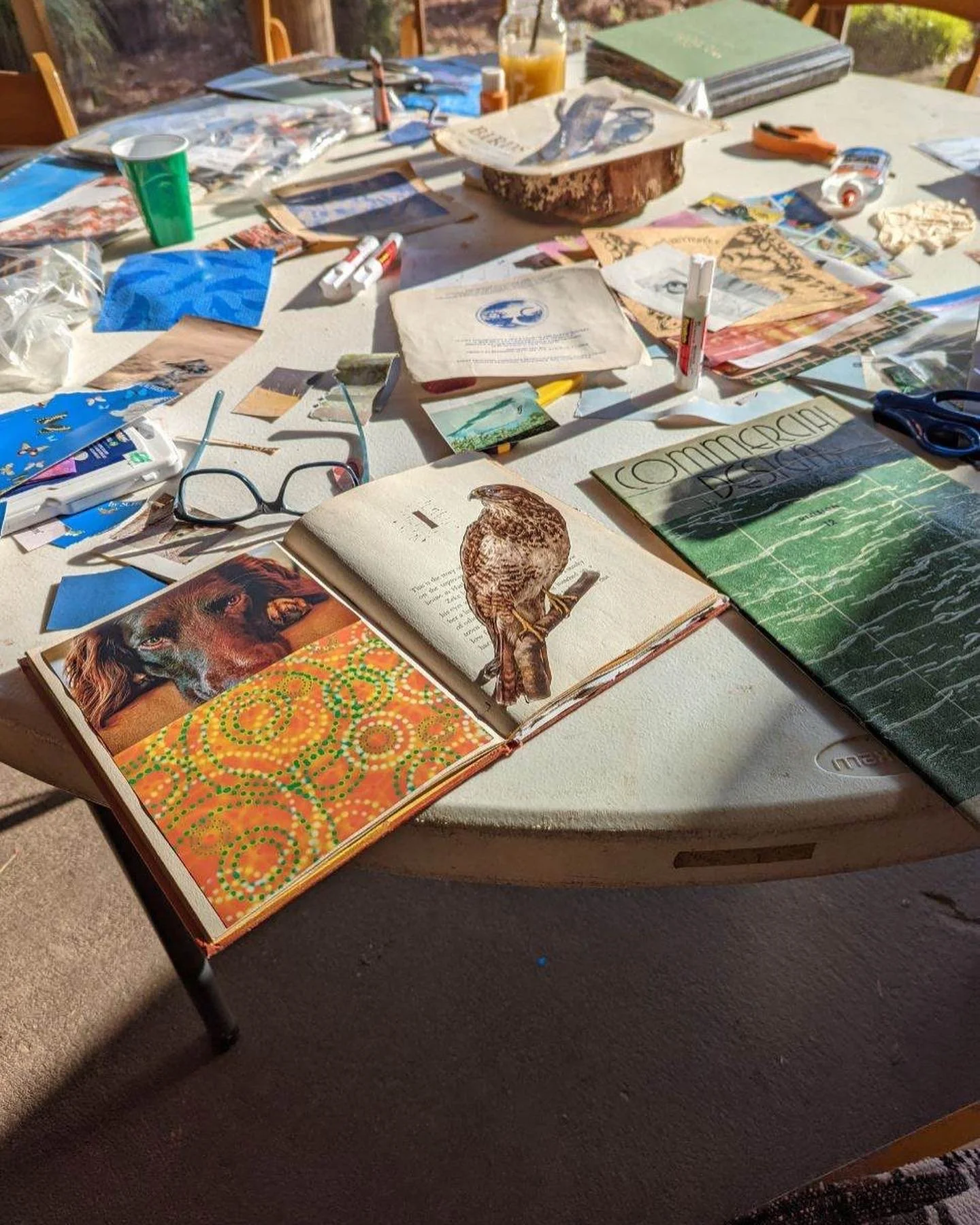 A cluttered table with magazines, books, scissors, glue, glasses, and various craft supplies, with sunlight casting shadows on the surface.
