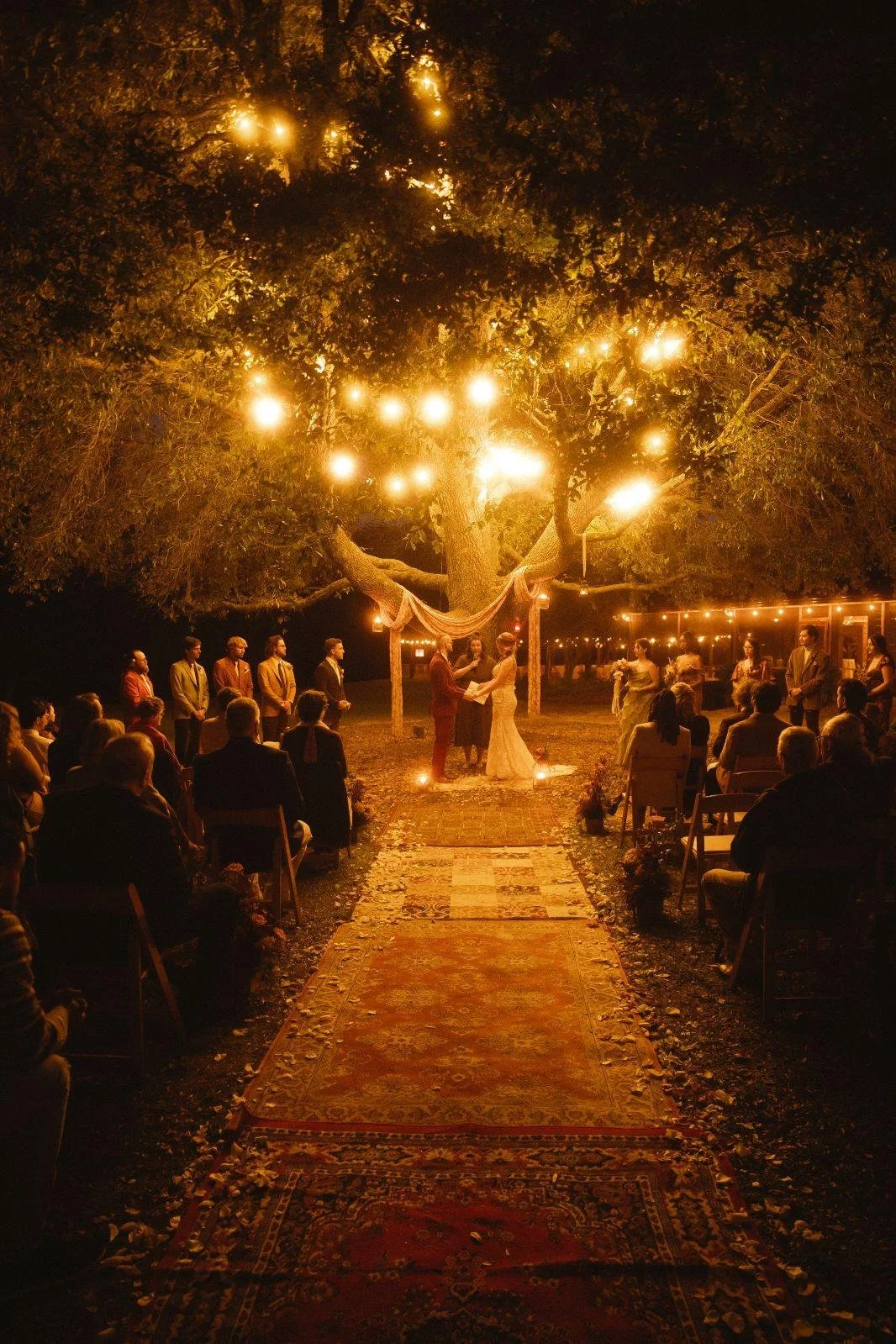 An outdoor wedding ceremony at night under a large lit tree with hanging string lights. The couple is exchanging vows at the altar, surrounded by seated guests and attendants.