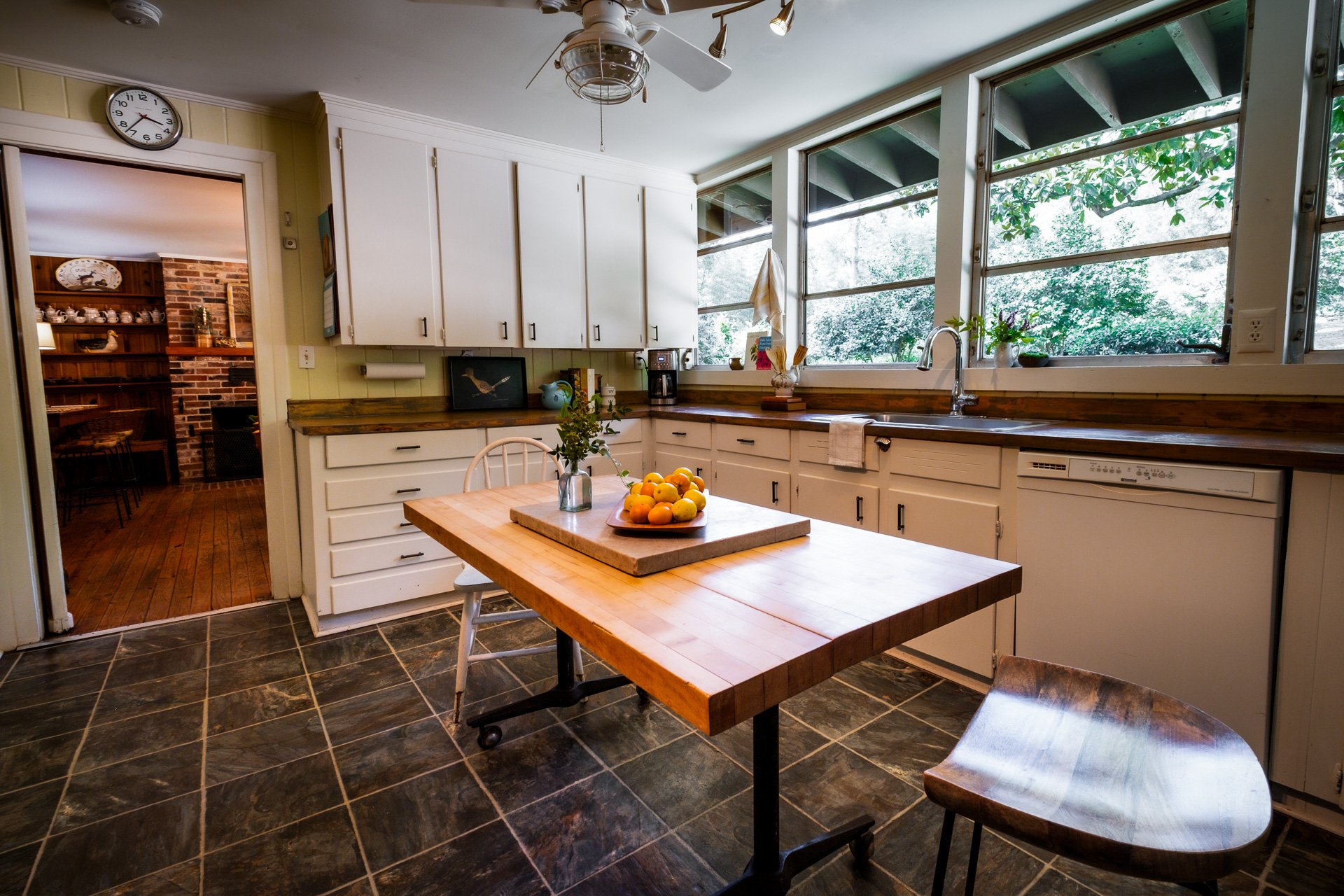 Kitchen with white cabinets, black tile floor, wooden table with a bowl of lemons, and large windows showing greenery outside.