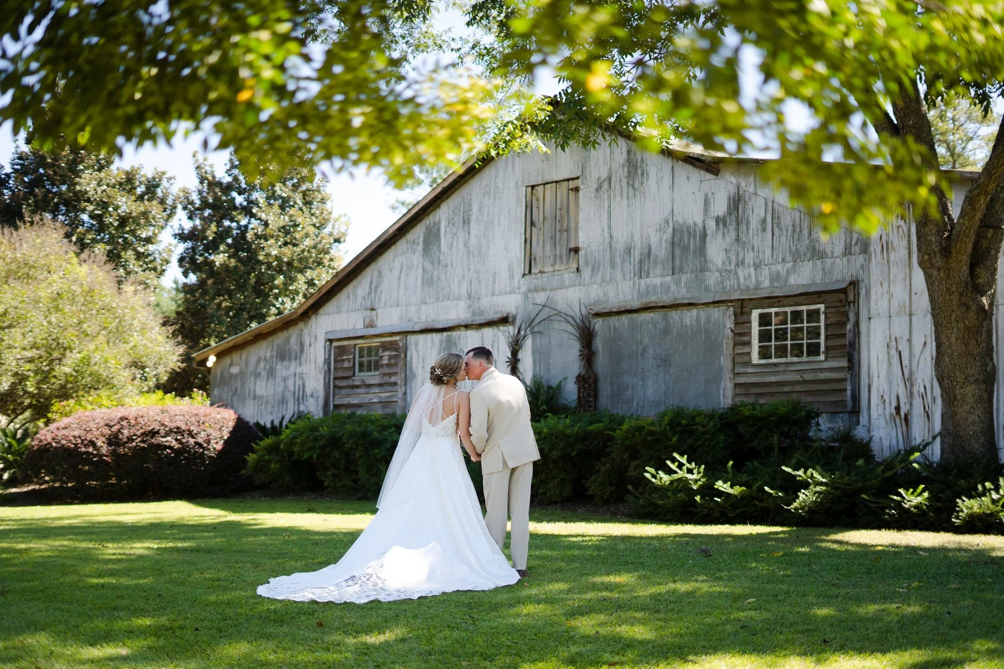 A bride and groom dressed in wedding attire sharing a kiss outdoors on a sunny day, with a rustic barn and trees in the background.