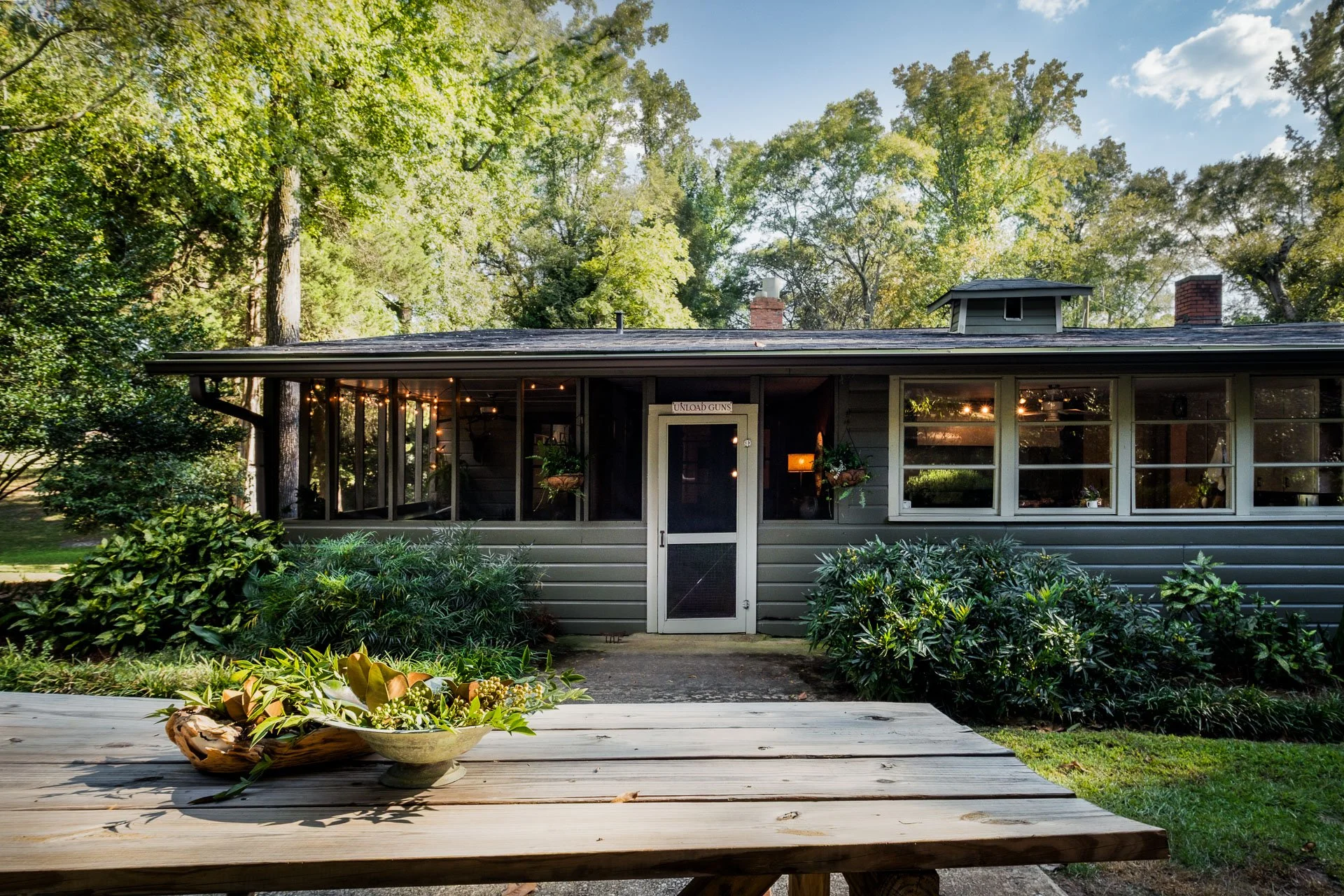 Front view of a house with a screened porch, wooden siding, and lush green bushes surrounding it. There are string lights visible inside the porch, and a sign above the door that says 'UNLOAD GUNS.' A wooden picnic table with a centerpiece of plants is in the foreground, with tall trees and a blue sky in the background.