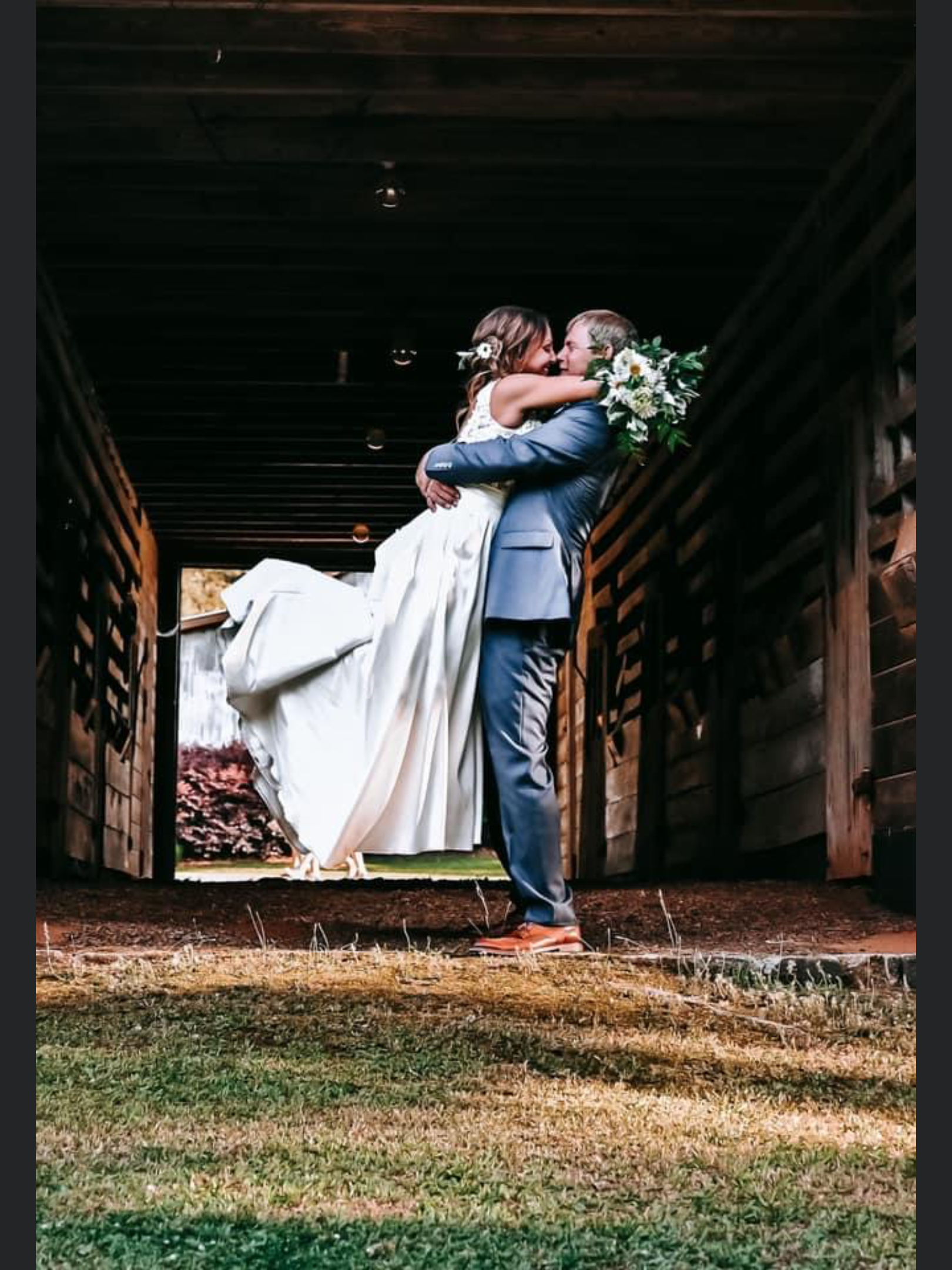 A bride in a white dress being lifted by a groom in a suit, holding a bouquet of flowers, inside a wooden structure.
