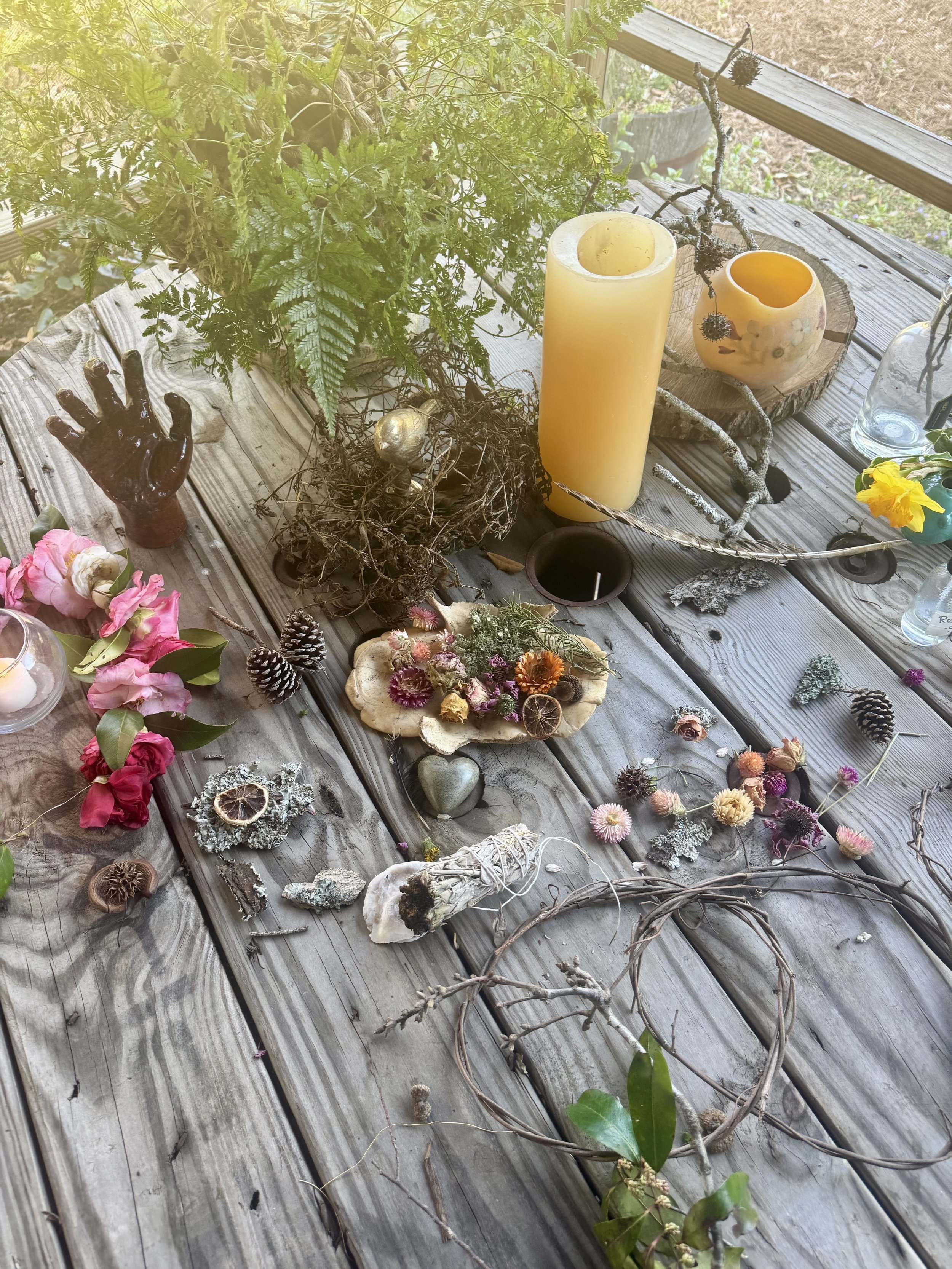A collection of decorative objects on a wooden table, including candles, dried flowers, pinecones, a black hand sculpture, and natural elements like leaves and branches, with greenery and outdoor scenery in the background.