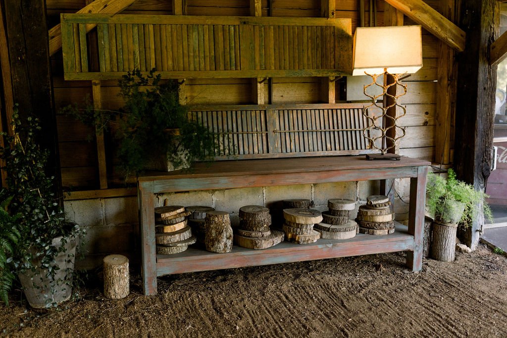 A rustic wooden table with various cut logs arranged on the lower shelf, set against a wooden wall inside a barn-like structure. There are plants on either side of the table and a decorative lamp on the right side of the table.