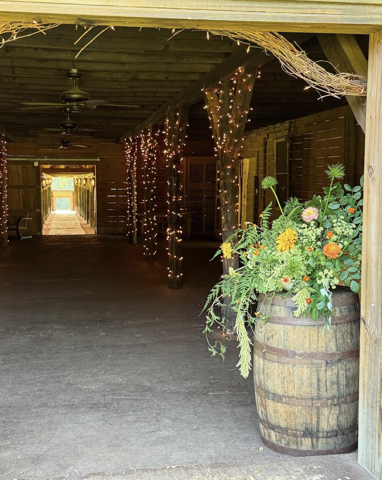 A rustic indoor space with wooden walls and ceiling, decorated with string lights, containing a large wooden barrel filled with colorful flowers, and a doorway leading outside.