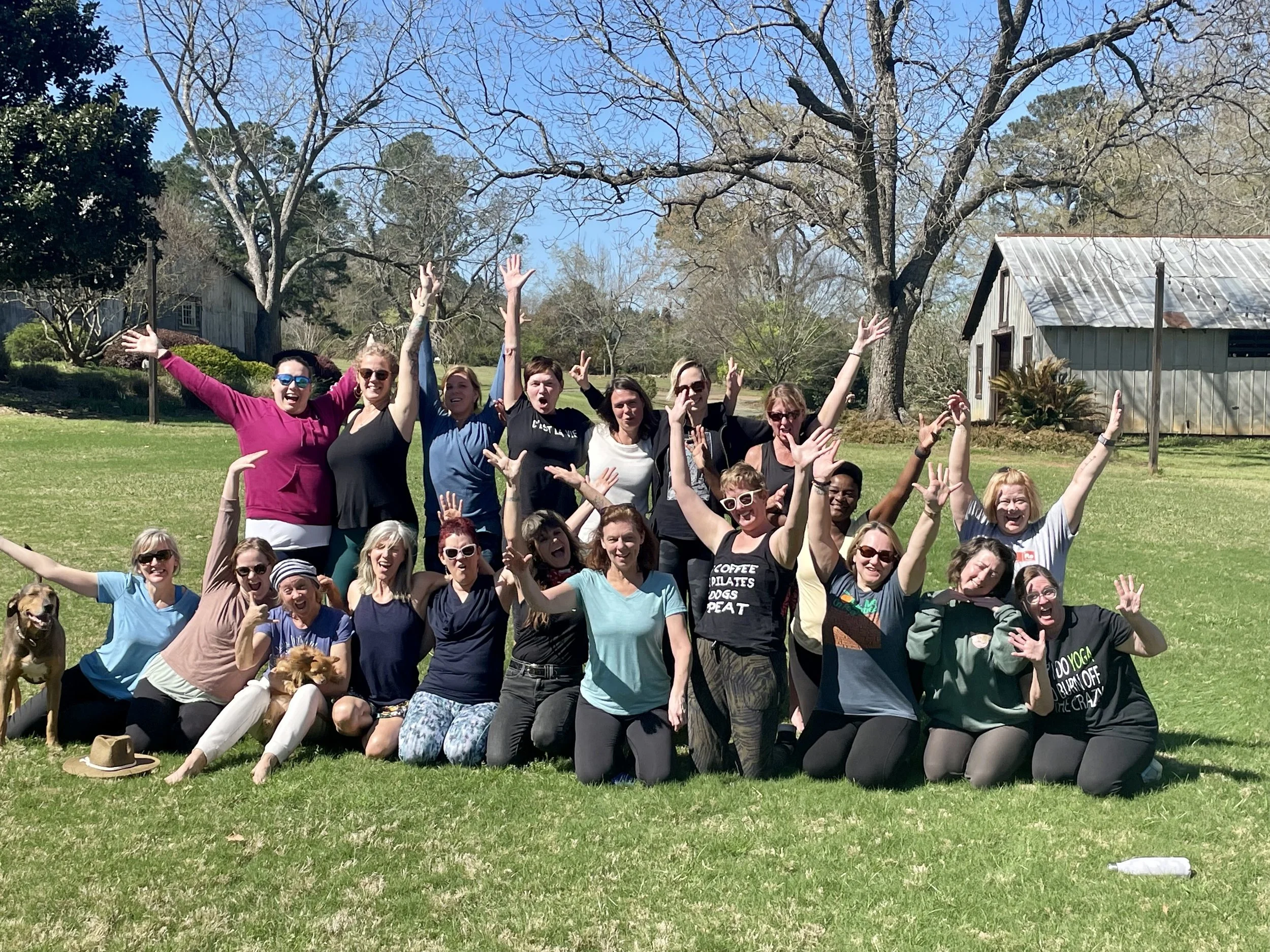 Group of smiling women and two dogs posing outdoors on a grassy field, with trees and a rustic barn in the background, celebrating and having fun.