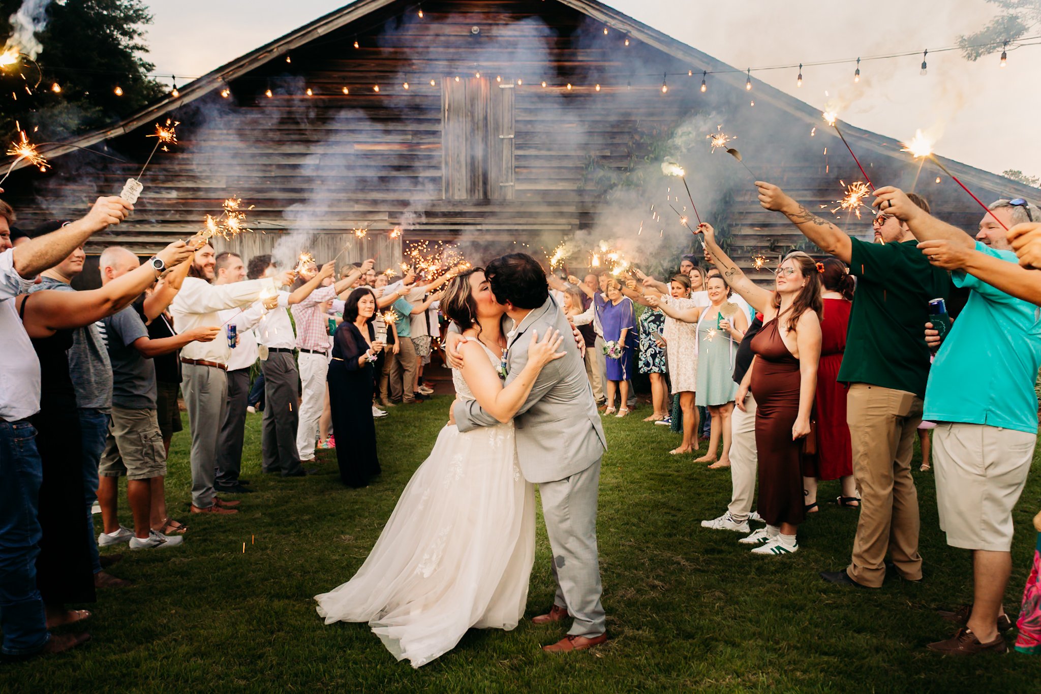 A newlywed couple sharing a kiss and kiss in the middle of a large group of friends and family holding sparklers during an outdoor wedding celebration at sunset.