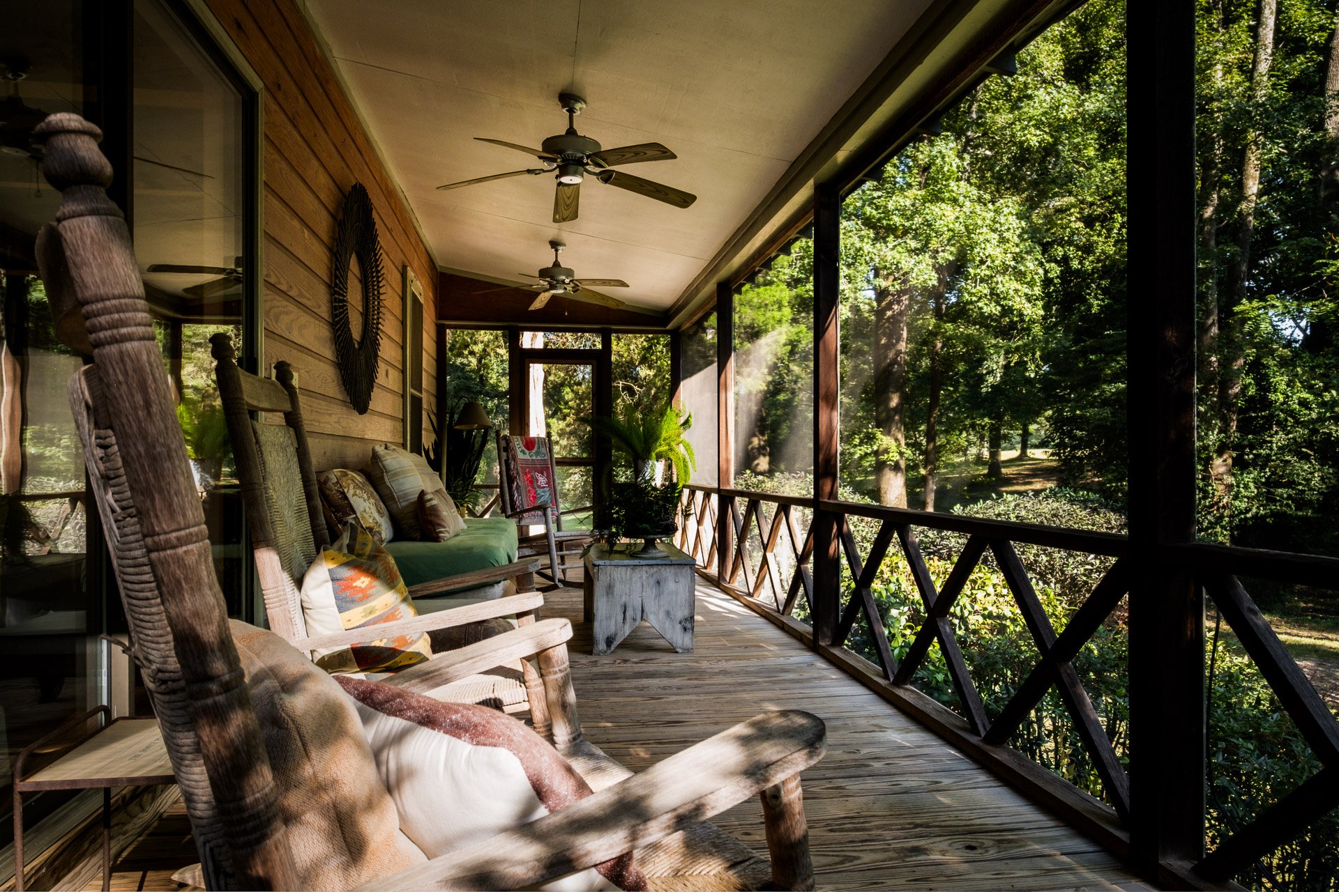 A spacious screened porch with wooden flooring and ceiling fans, furnished with rustic chairs, a sofa, and a small table, overlooking a lush green forest.