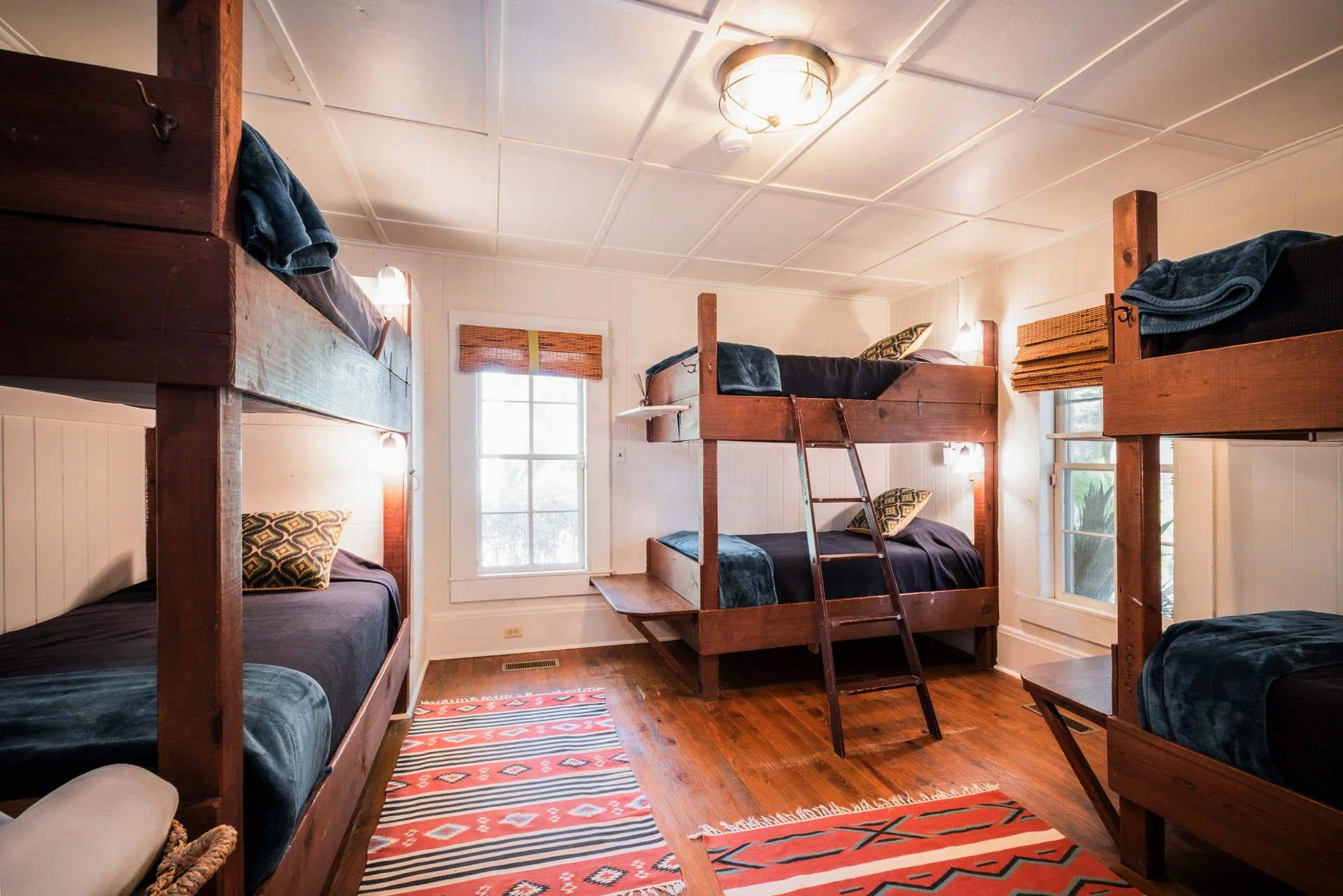 A bedroom with four wooden bunk beds, two on each side of the room. Each bed has dark bedding and a pillow with a patterned pillowcase. There are two windows with blinds, and a ceiling light fixture. The floor is wooden with three colorful area rugs.
