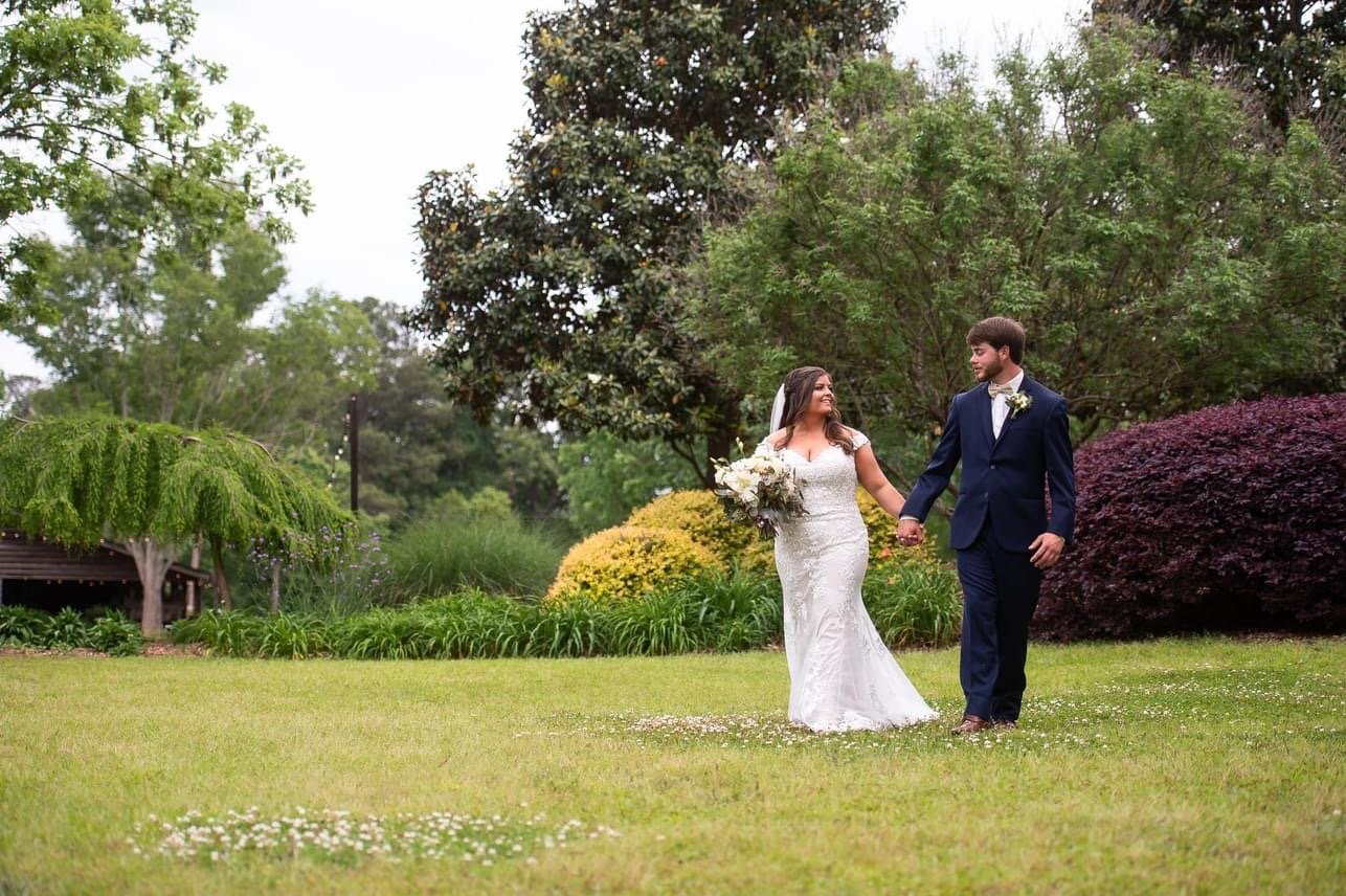 A bride and groom holding hands and walking in a lush garden with trees and colorful bushes.