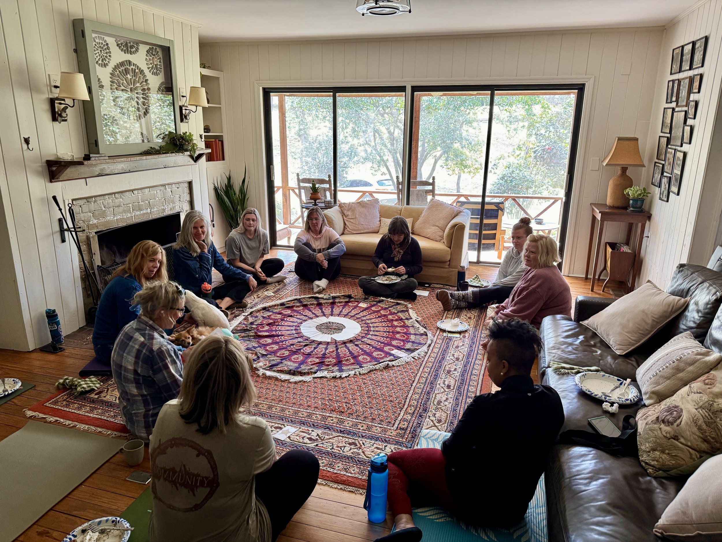 Group of people sitting on the floor and couches in a living room during a gathering or meeting, with a large sliding glass door leading to an outdoor balcony.