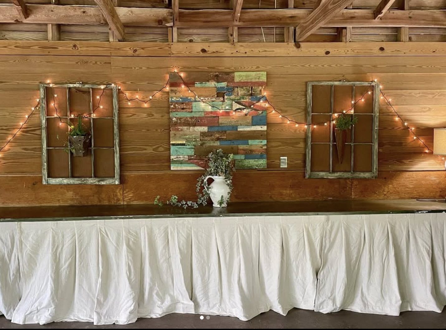 Rustic wooden wall with two window frames and string lights, decorated with potted plants, a colorful wooden art piece, and a white vase with greenery, with a table covered in a white cloth in front.