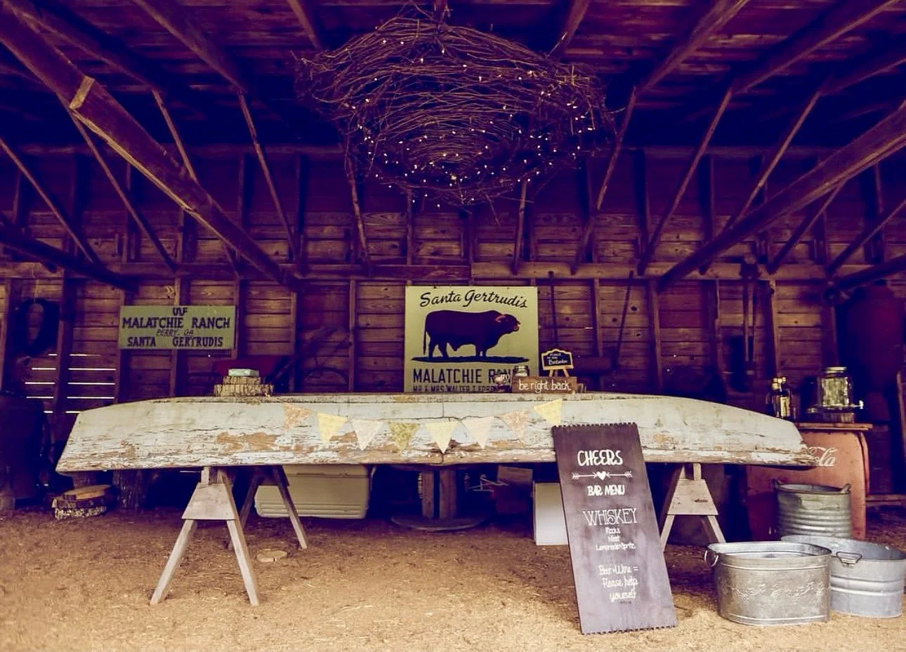 Inside a rustic barn, a dark wooden table is decorated with a banner, and signs advertise Santa Gertrudis cattle, whiskey, and a barbeque menu. The walls and ceiling are made of wooden planks and beams, with a decorative light fixture hanging from th