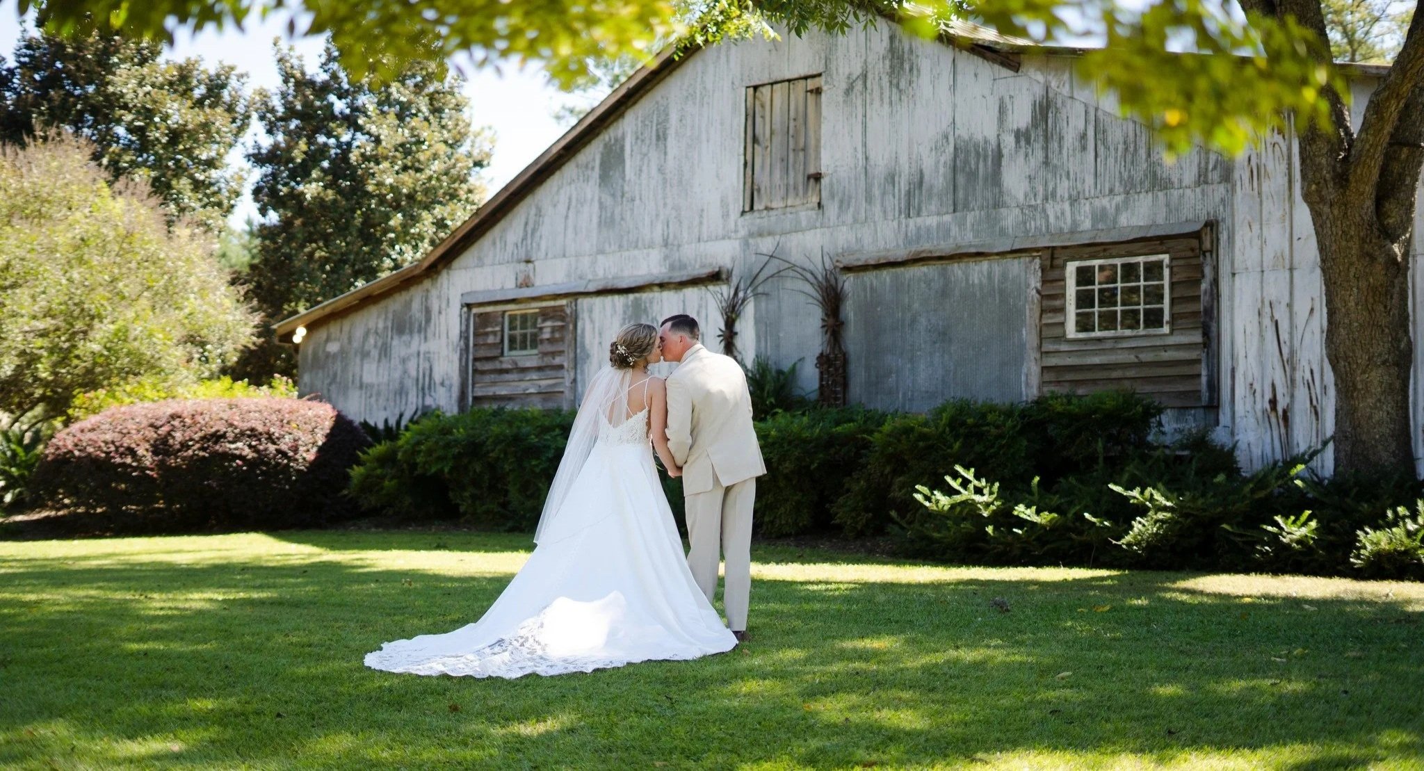 A bride and groom share a kiss outdoors in front of an old wooden barn, surrounded by green grass, bushes, and trees on a sunny day.