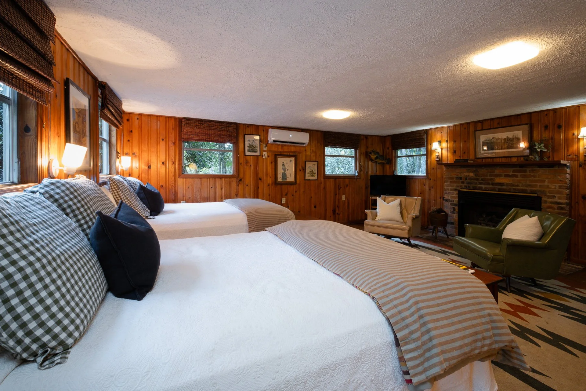 A cozy bedroom with wood-paneled walls, featuring two beds with checkered and plain pillow arrangements, a fireplace, a green armchair, a beige armchair, a television, and multiple windows showing greenery outside.