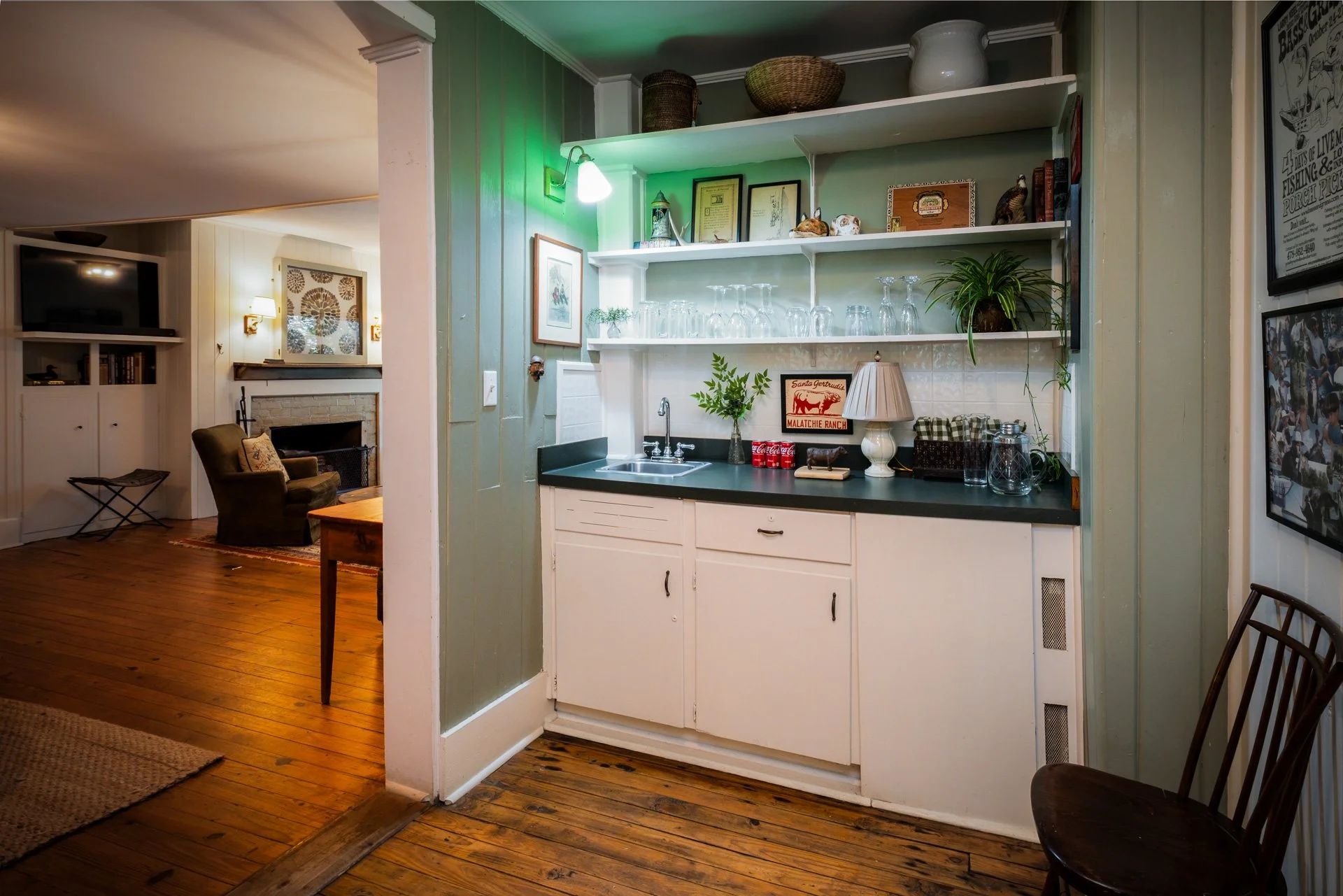 A small kitchenette with white cabinets, green countertops, open shelves holding glasses and decorative items, and a small sink, near a cozy living room with wooden flooring, armchair, and fireplace.