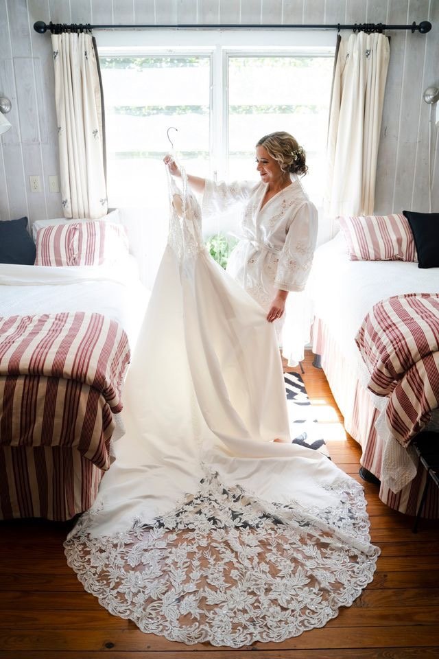 A woman in a white bathrobe smiling while holding a wedding dress in a bedroom with natural light, two beds with striped bedding, and a window behind her.