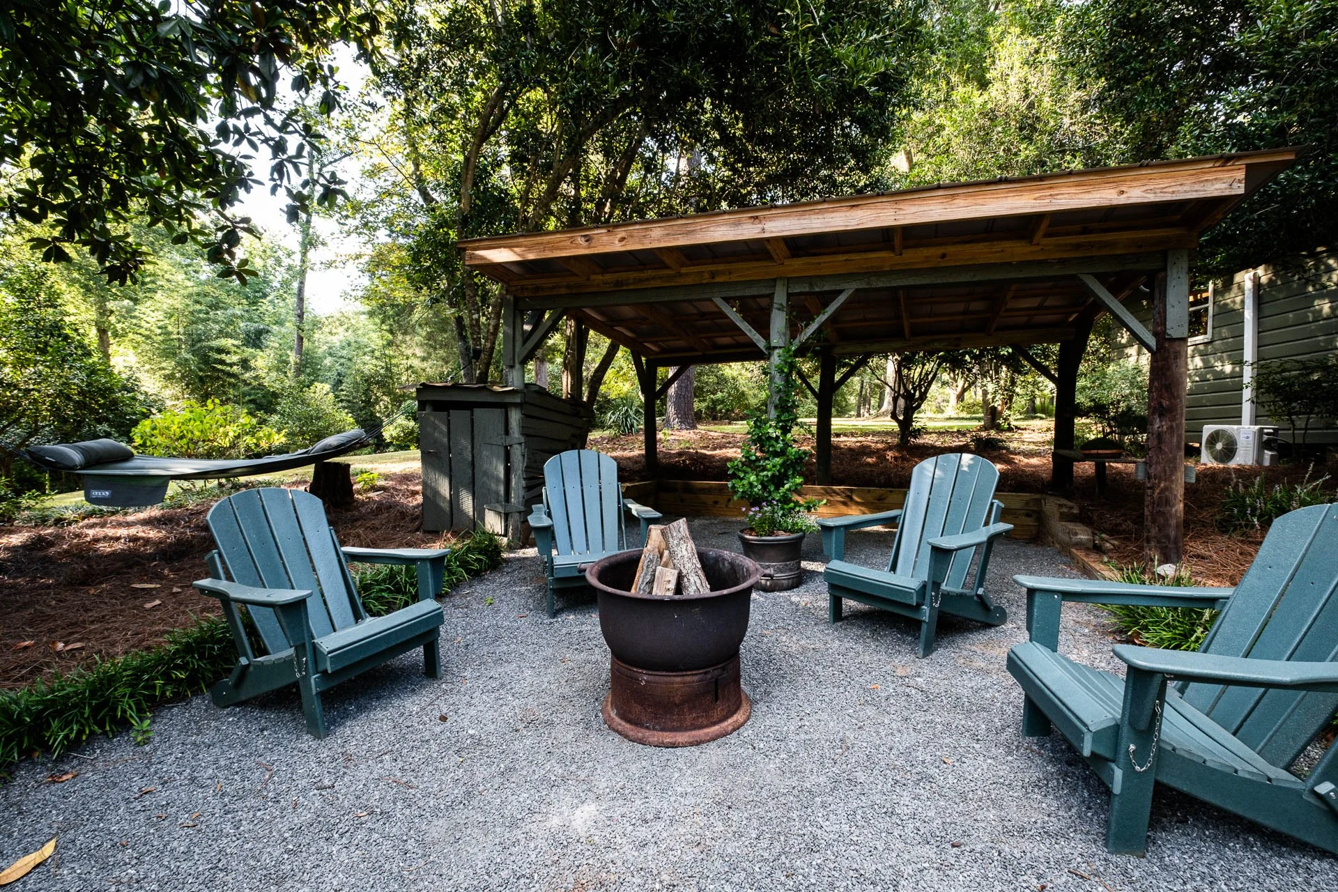 Outdoor seating area with four blue Adirondack chairs arranged around a fire pit, under a wooden shelter, surrounded by trees and greenery.